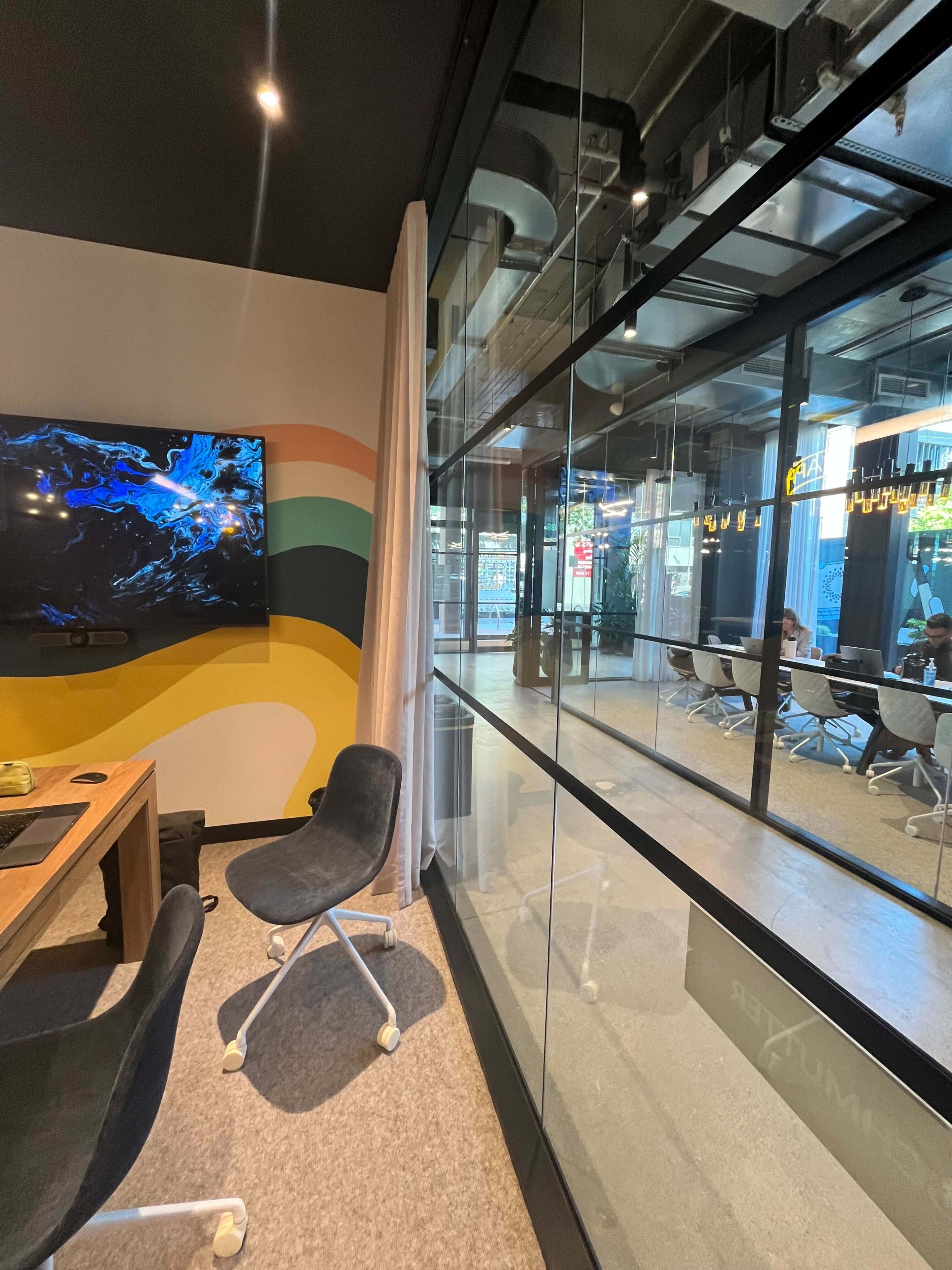 A modern office space features a glass-walled meeting room with a view of people sitting at a table, and a colorful mural on one wall.