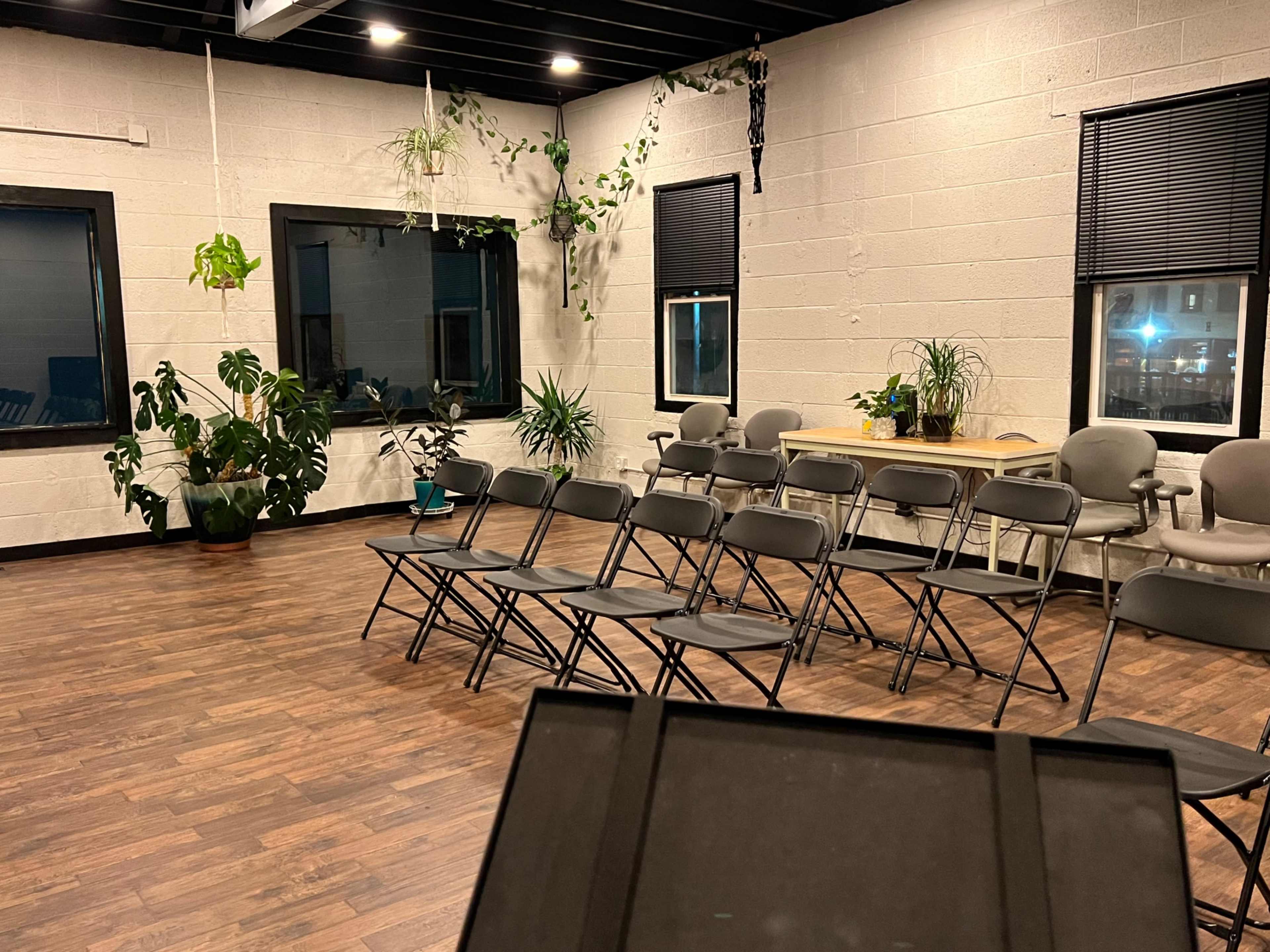 A spacious room with folding chairs arranged in rows, plants positioned near windows, and a table against the wall.