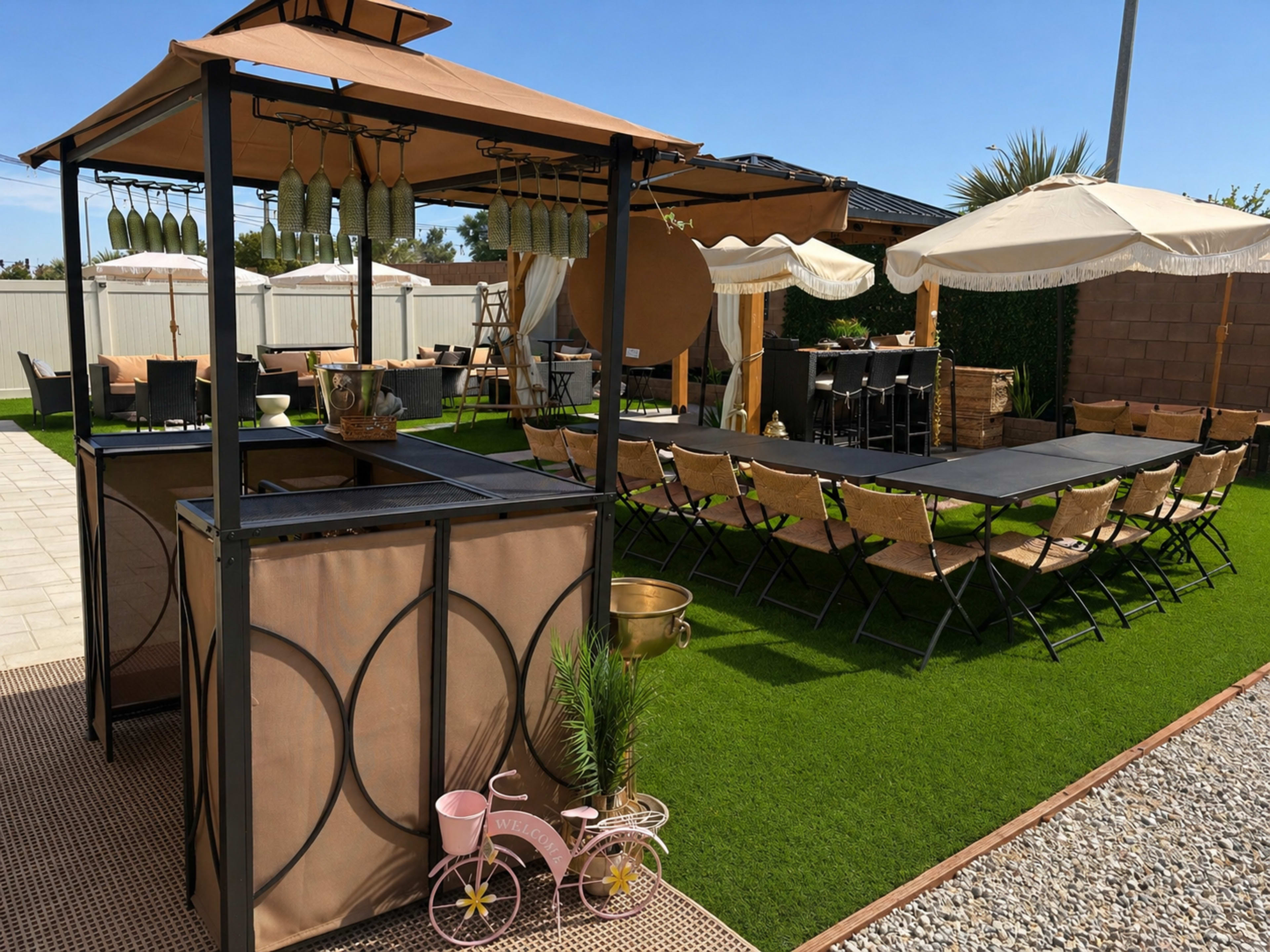The image shows an outdoor patio area featuring a gazebo with hanging decorations, a bar setup, multiple seating arrangements, and several umbrellas on green grass.