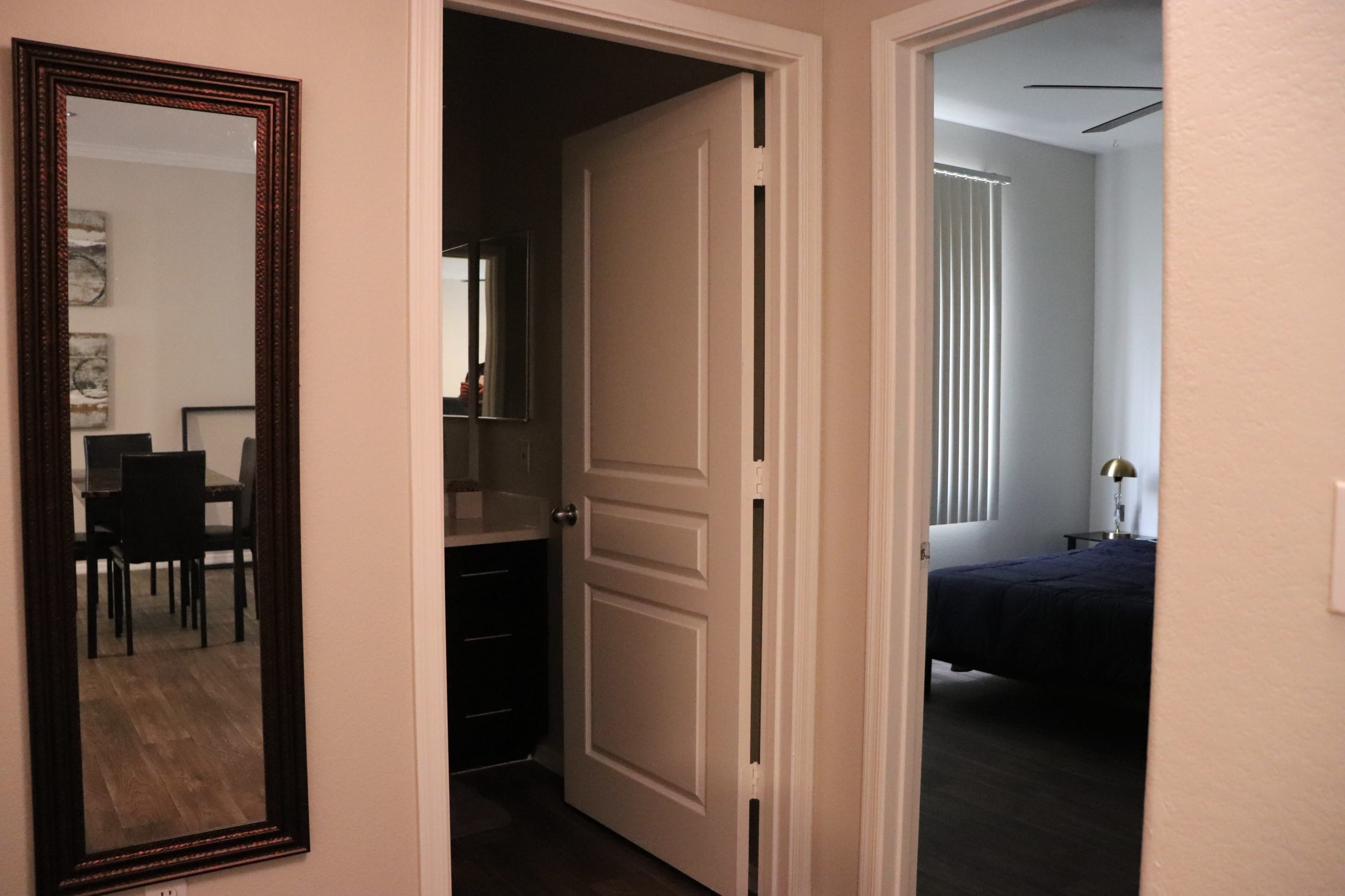 The image shows a hallway leading to two separate rooms, with a mirror on the left and a view into a dining area and a bedroom.