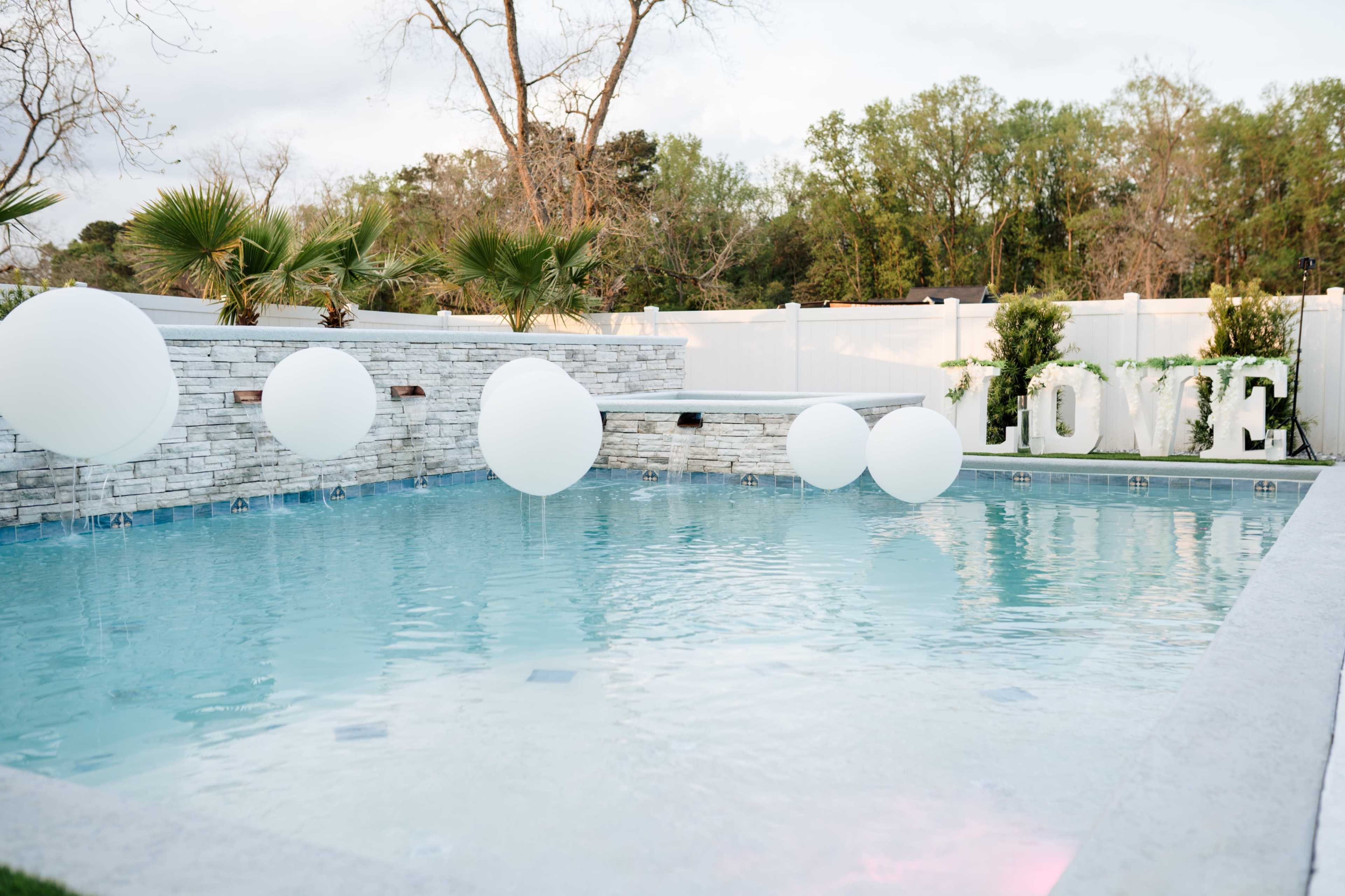 A swimming pool features large white balloons floating above the water, with greenery and a stone wall in the background displaying the word "LOVE."