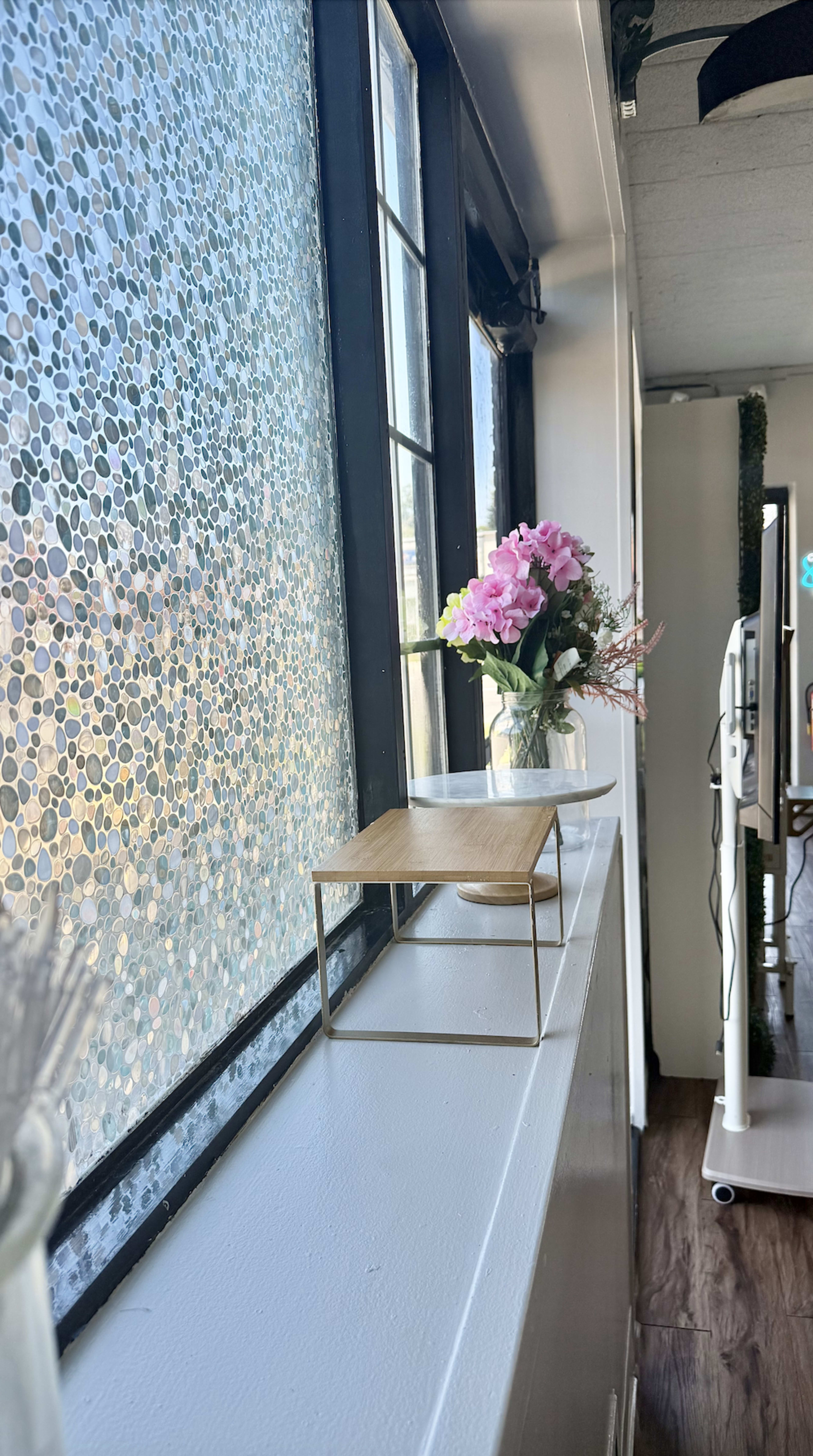 A small wooden table sits on a windowsill next to a vase of pink flowers, with a patterned window allowing light to filter through.