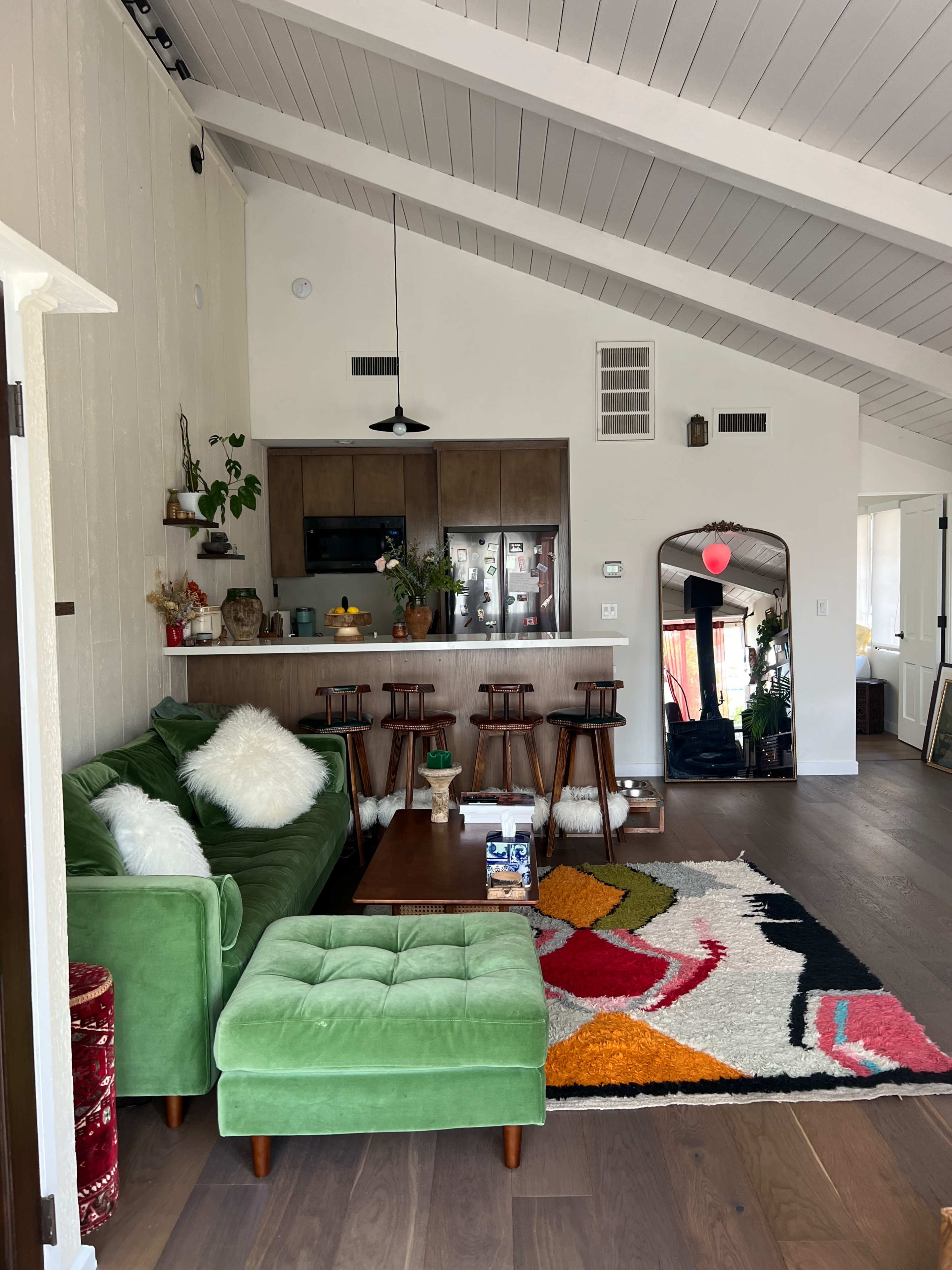 The image shows a brightly lit living area featuring a green sofa, a tufted ottoman, a wooden coffee table, and a kitchen bar with stools in the background.