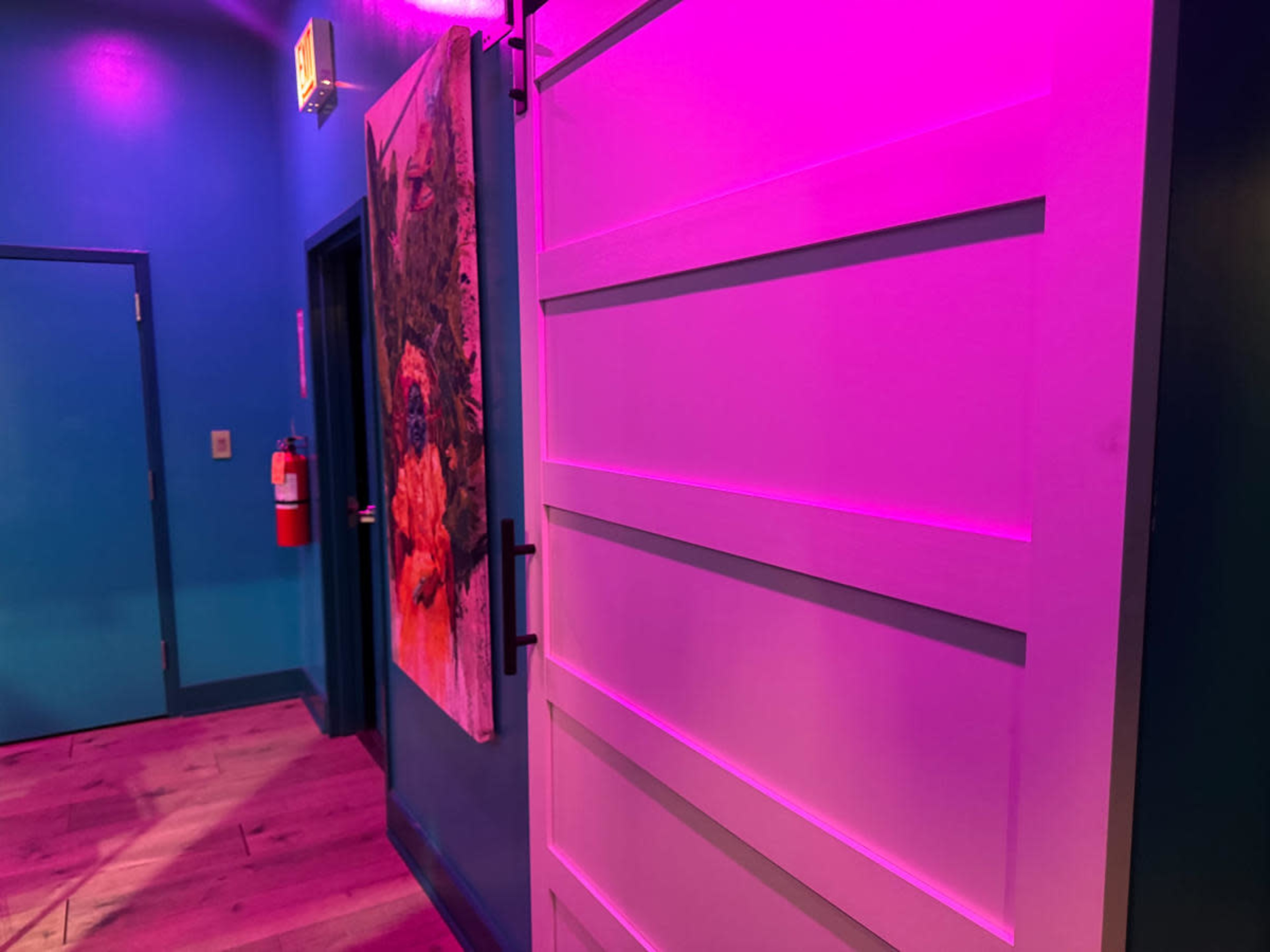 The image shows a brightly lit hallway with a white door on the right and a colorful artwork on the left, all illuminated by purple and blue lighting.