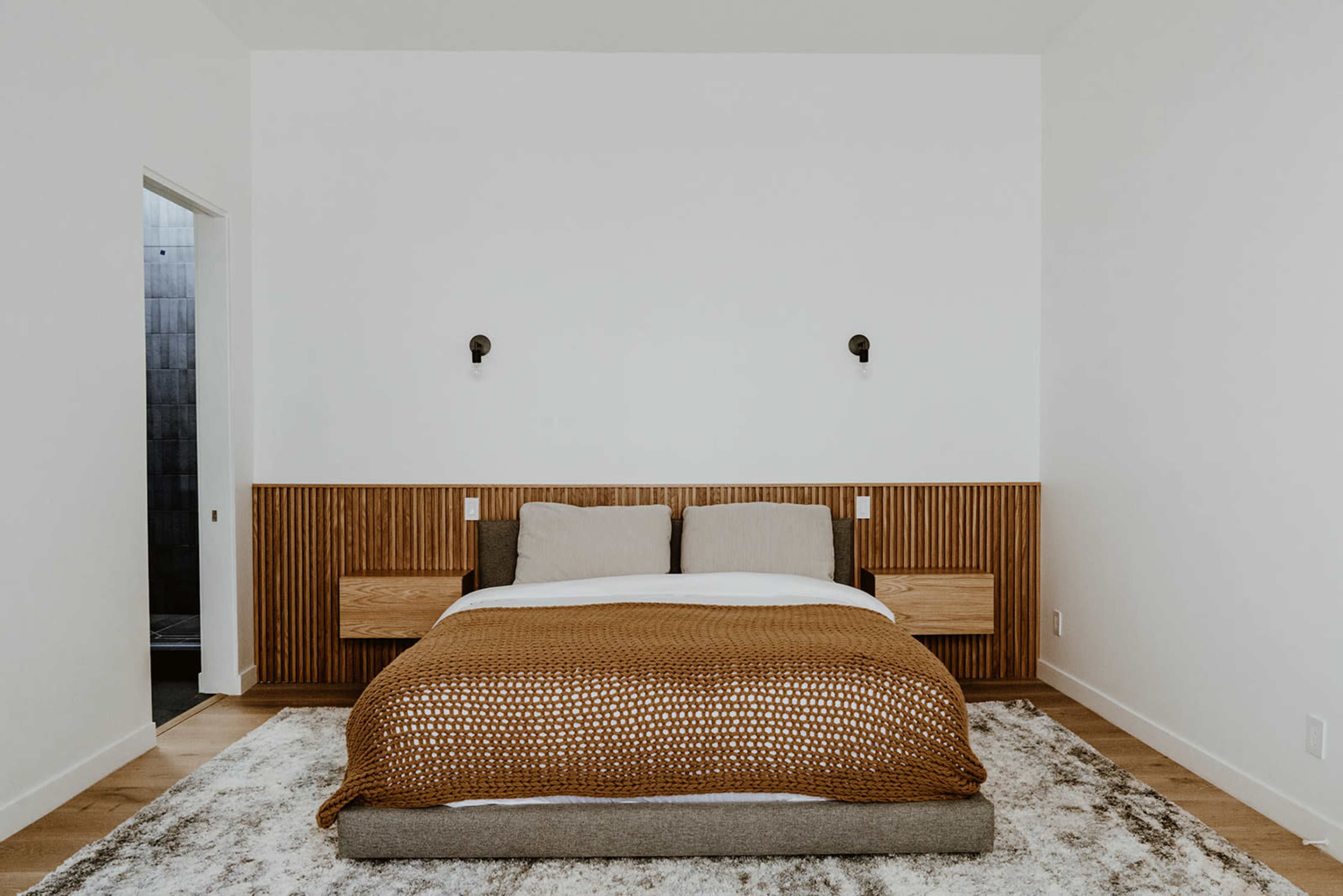 The image shows a tidy bedroom featuring a bed with a textured brown throw and two bedside lamps attached to a wooden accent wall.