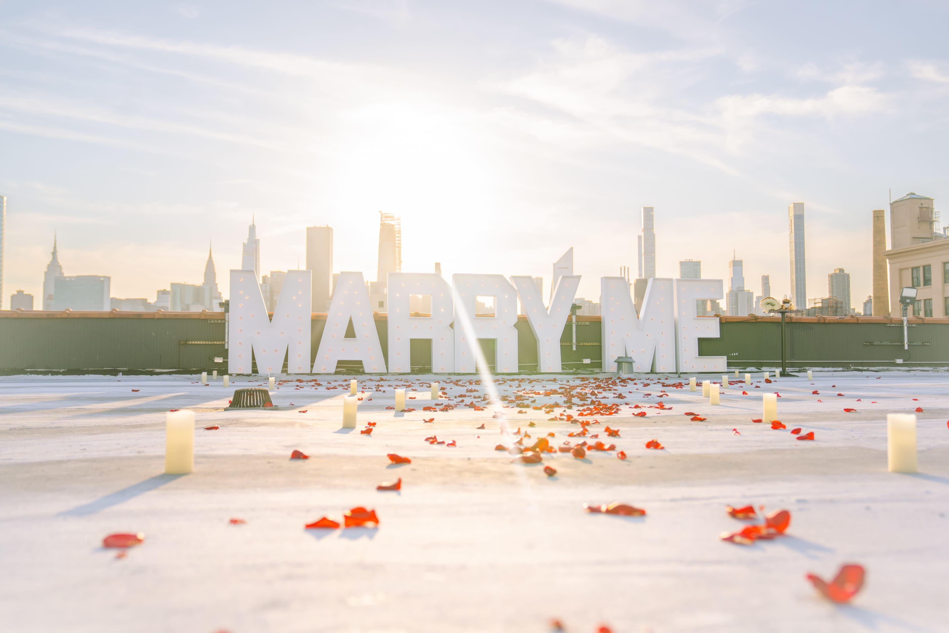 The scene features large illuminated letters spelling "MARRY ME," surrounded by scattered rose petals and candles, with a skyline view in the background.