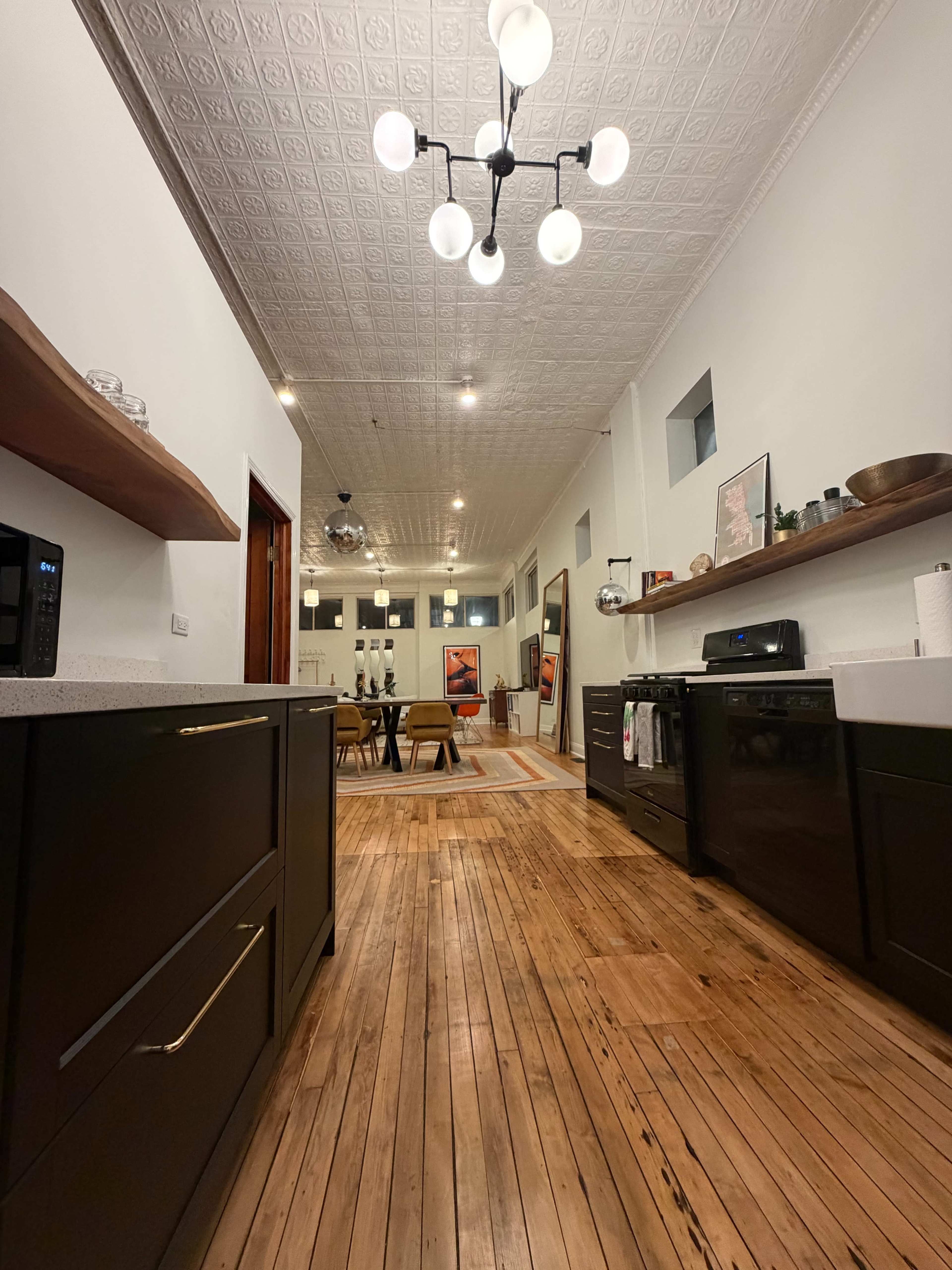 The image shows a modern kitchen with dark cabinets and a wooden floor, leading towards a dining area illuminated by a decorative ceiling light.