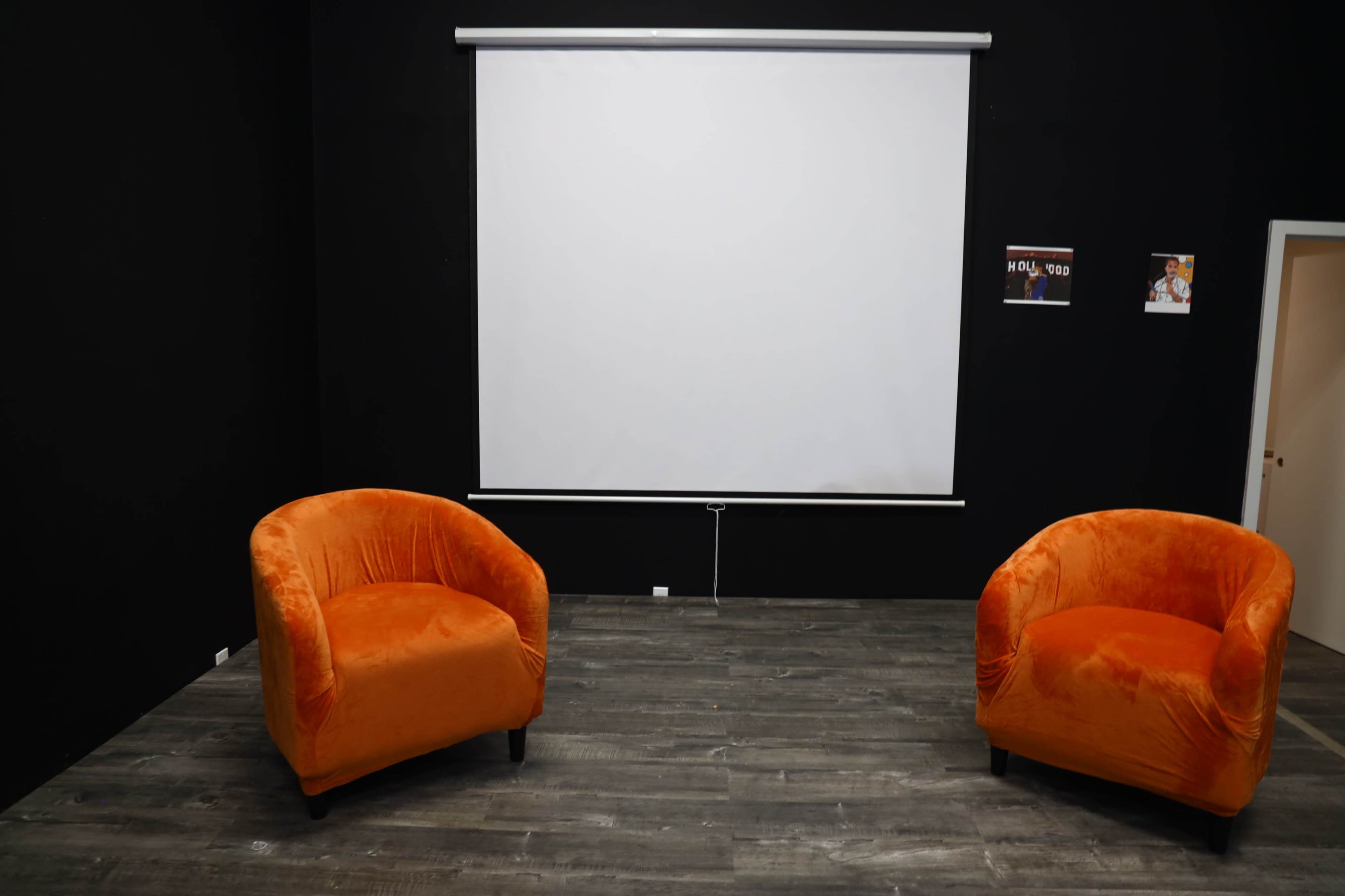 The image shows two orange armchairs facing a blank projection screen on a dark wall in a sparsely furnished room.