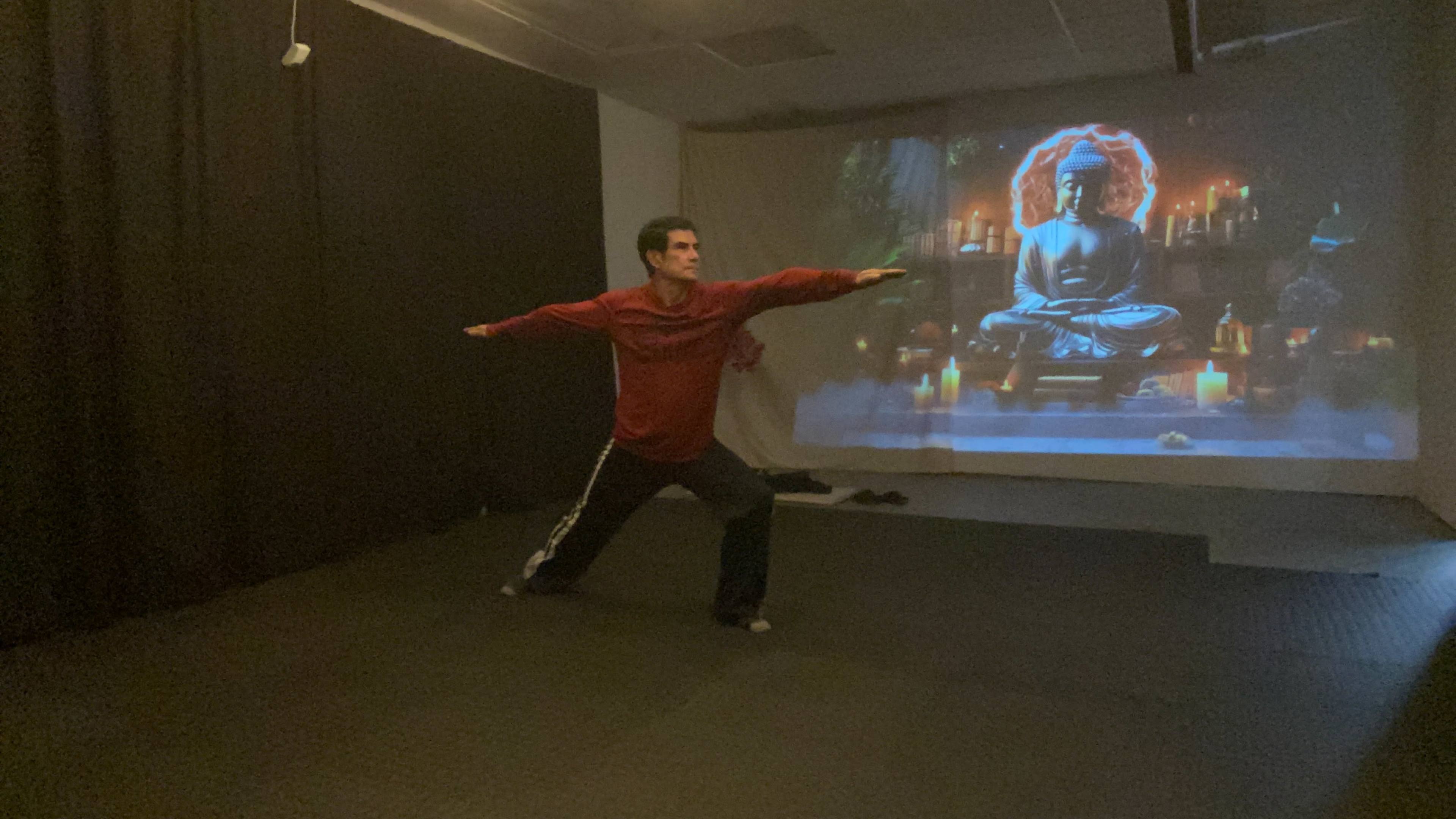 A person in athletic clothing performs a yoga pose in a dimly lit room with a projected image of a seated Buddha in the background.