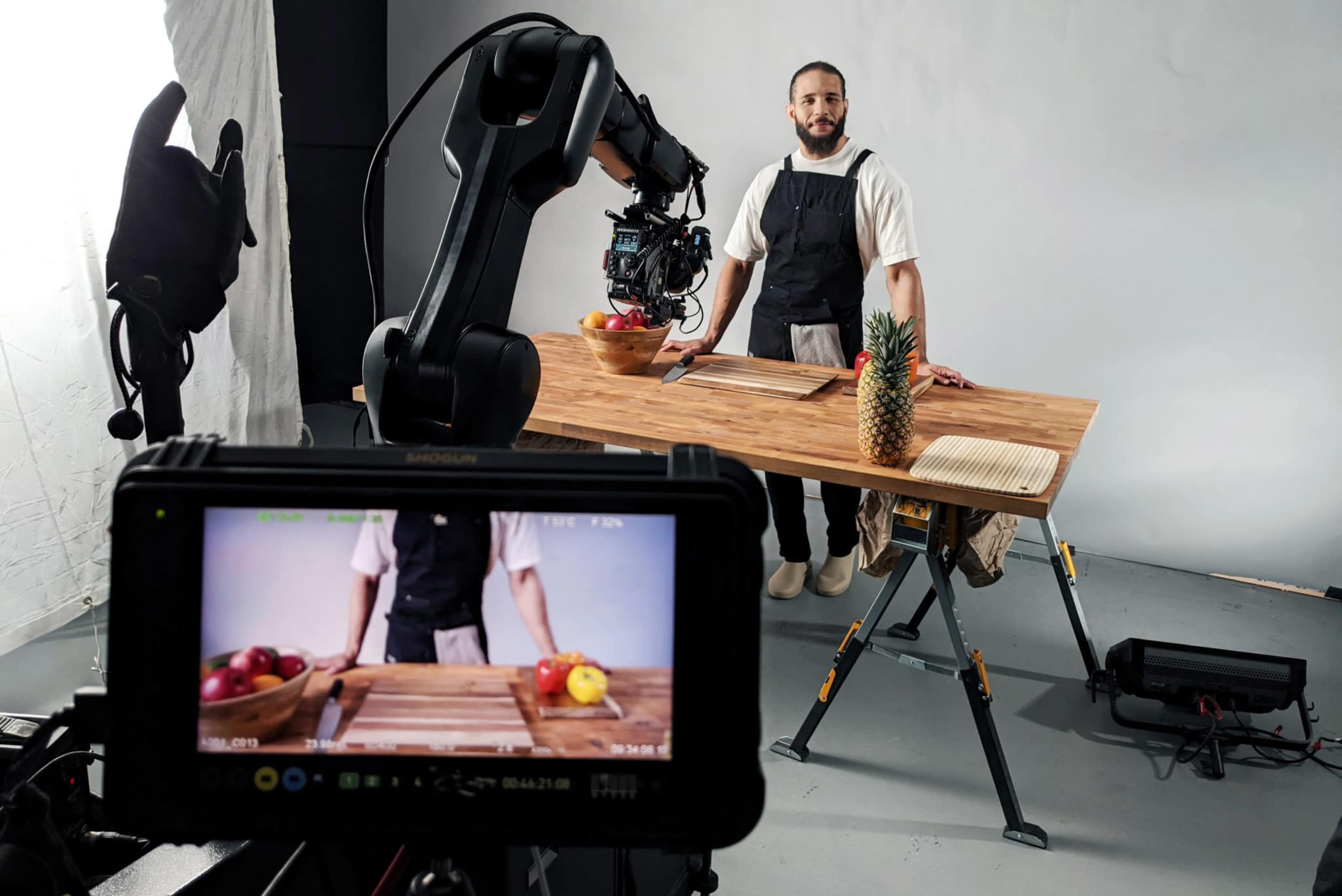 A man in an apron stands behind a wooden table with a bowl of fruits and a pineapple, while a camera on a tripod is focused on the scene.