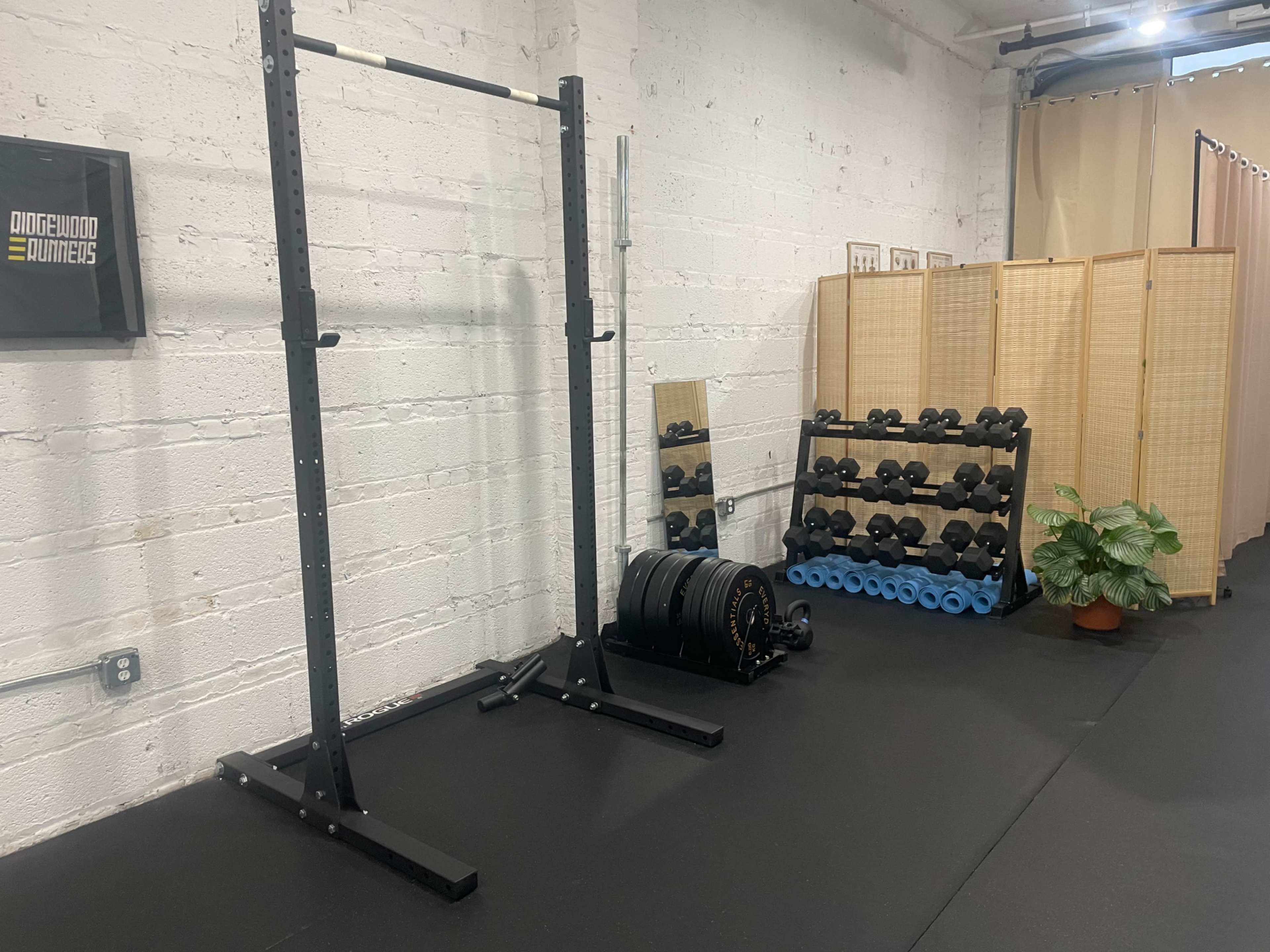 The image shows a home gym setup featuring a power rack, a mirror, a rack of dumbbells, and a storage area for weights in a well-lit room with black flooring.