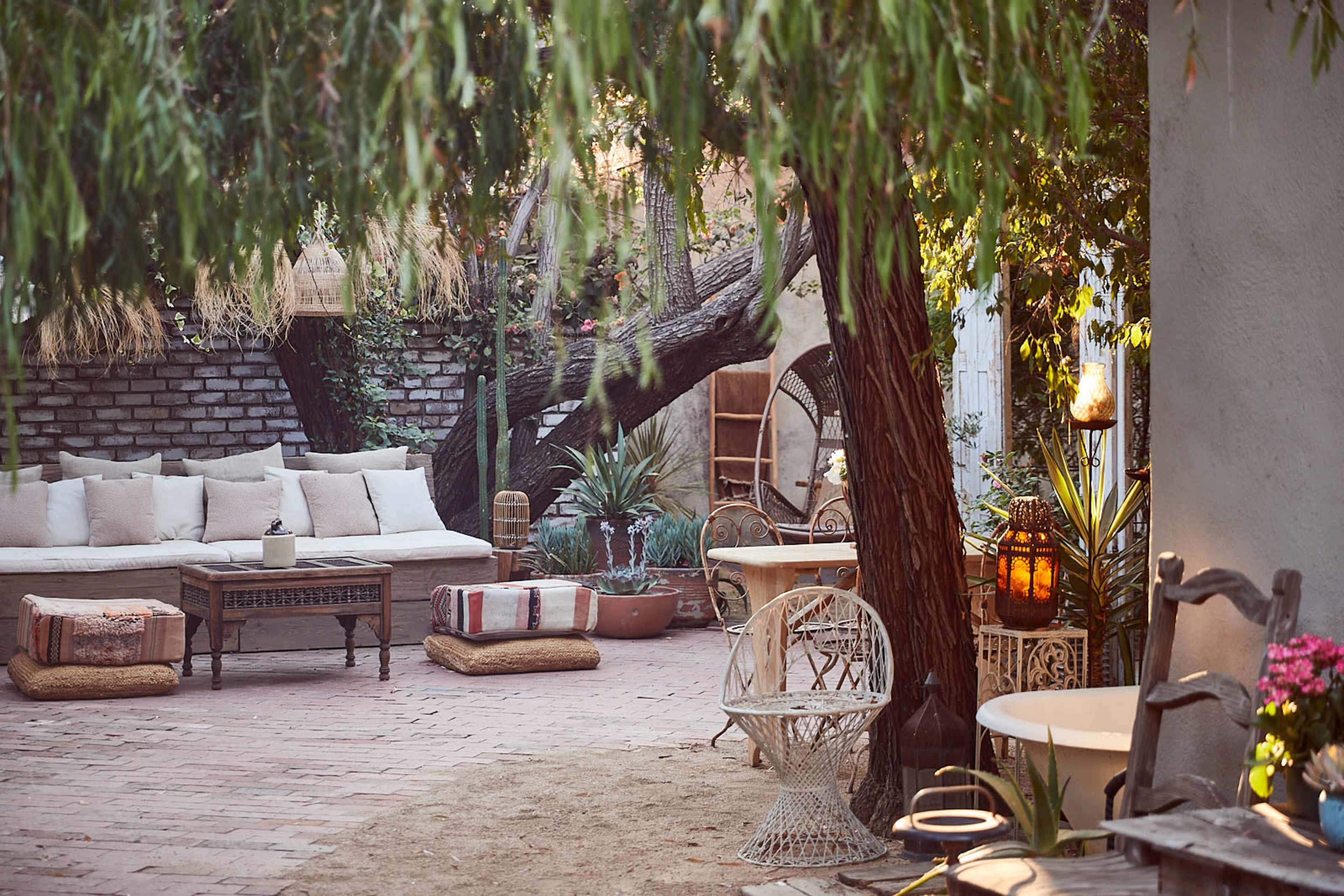 A cozy outdoor seating area features a sofa, wooden furniture, potted plants, and decorative lanterns beneath a large tree.