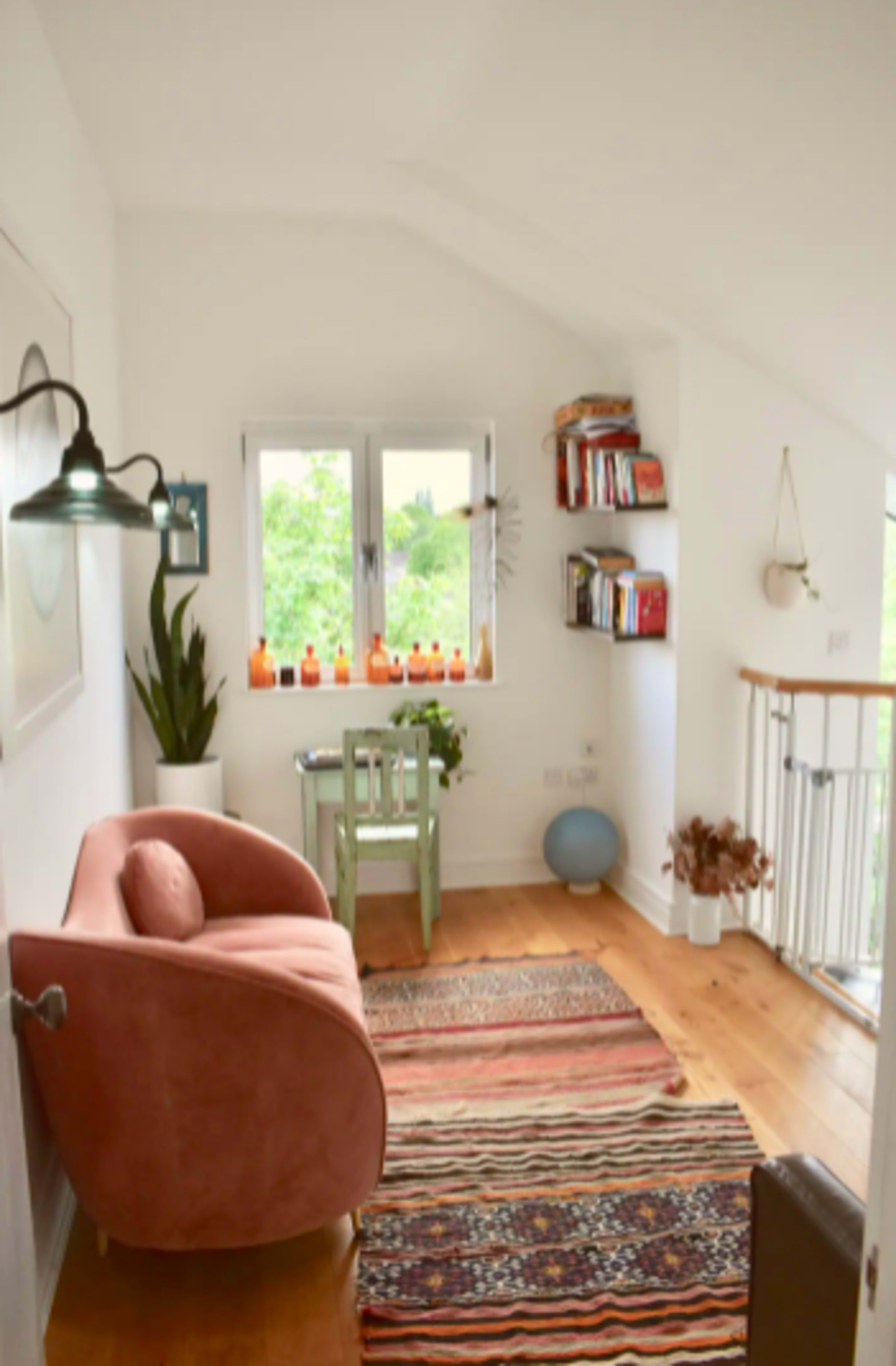 The image shows a bright, cozy corner of a room featuring a pink sofa, a small green table, and a collection of books displayed on a shelf.