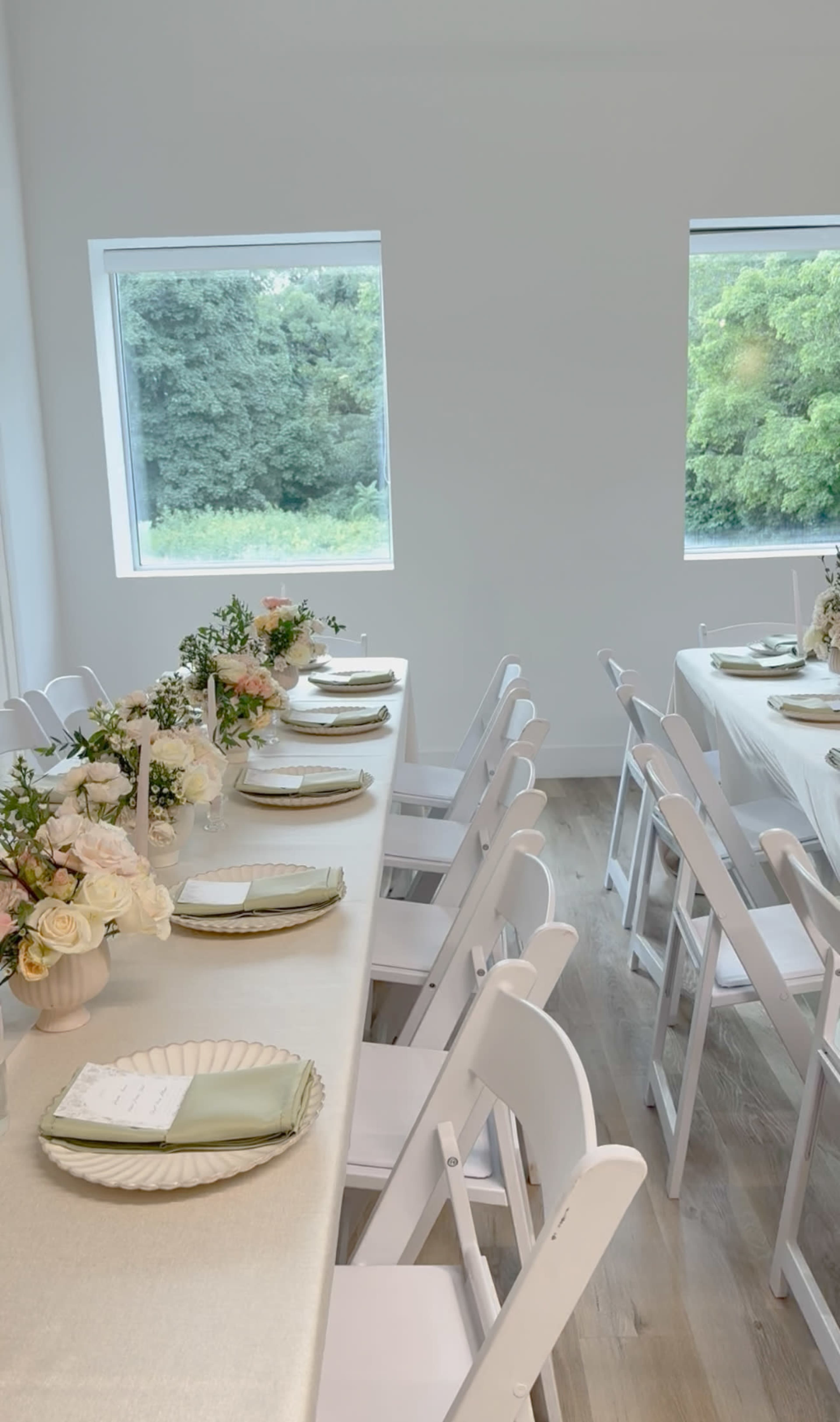 The image shows a dining setup with two long tables adorned with flowers and place settings, positioned in a bright room with large windows overlooking greenery.