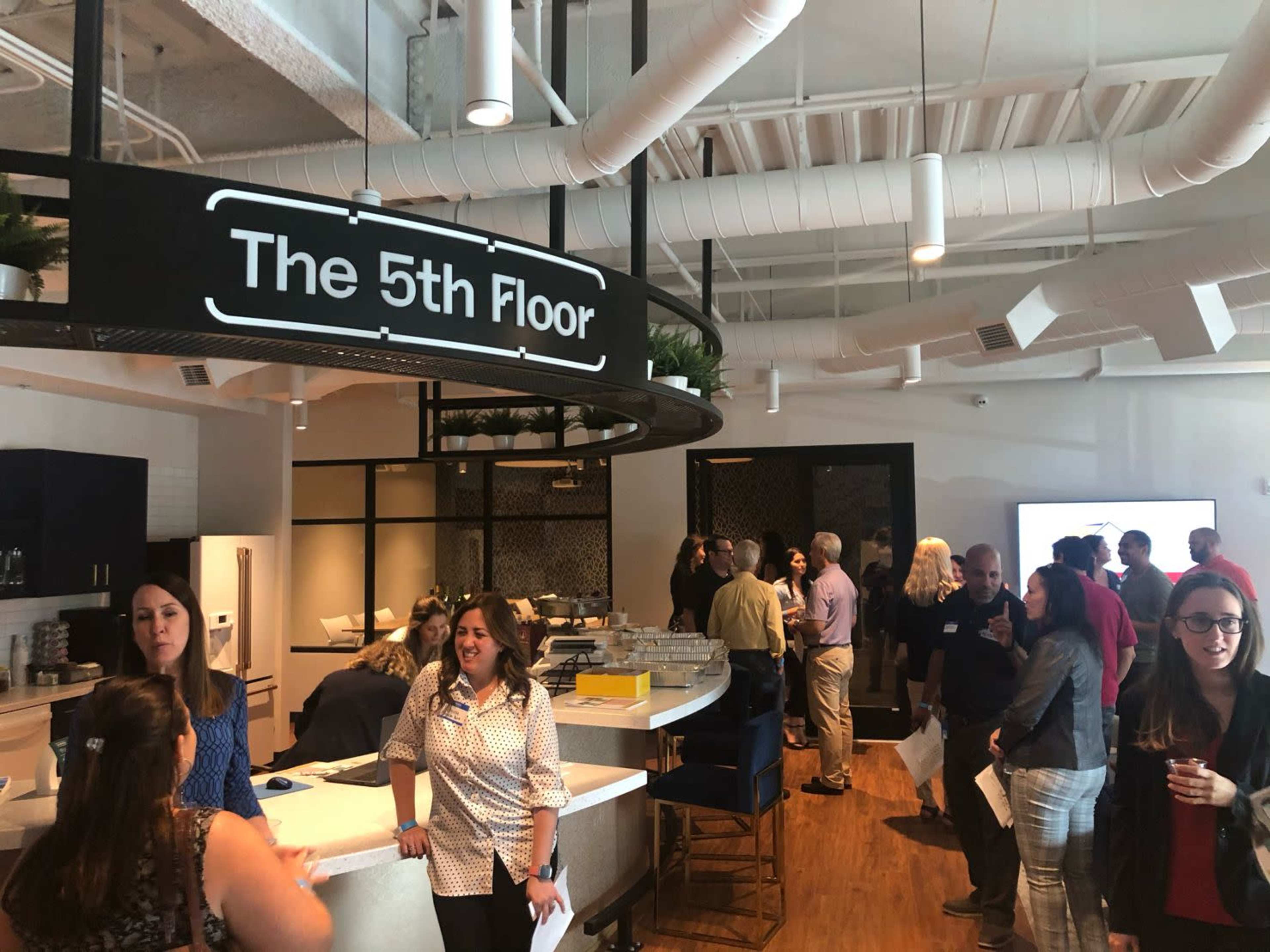 A group of people gathers in a modern open space featuring a large sign that reads "The 5th Floor" above a kitchen area.