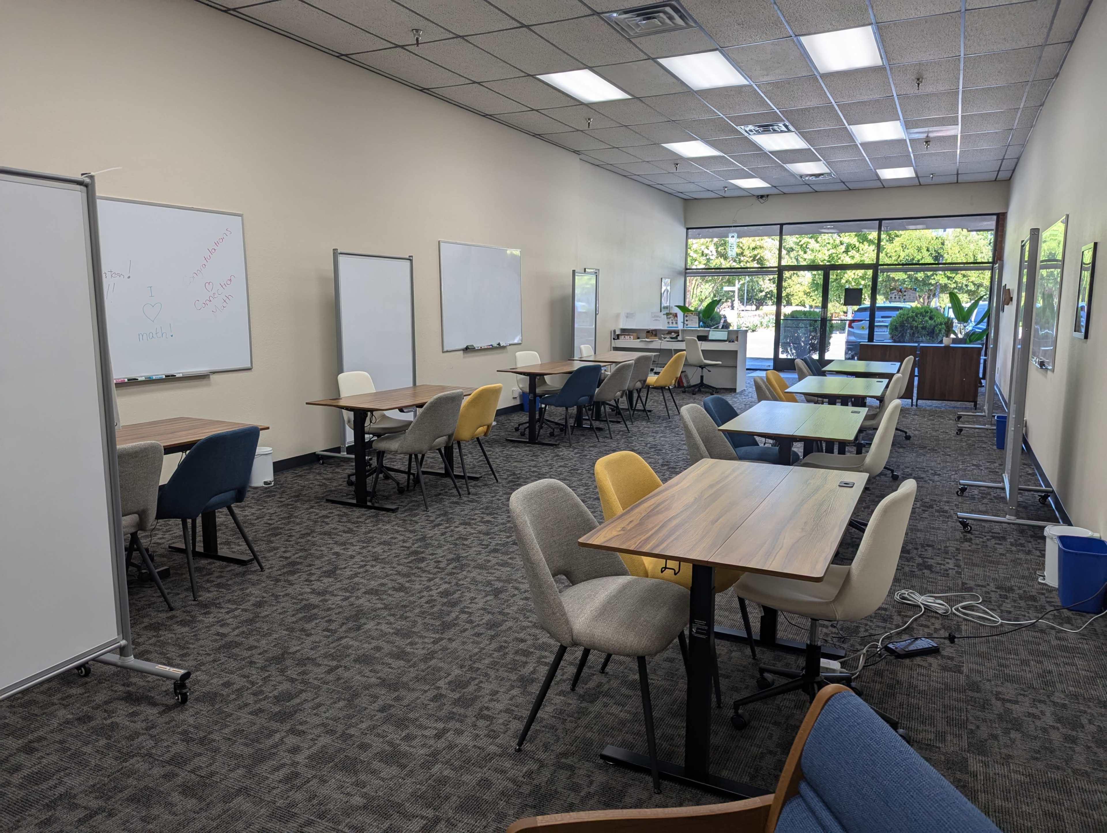 A spacious classroom or meeting room with several empty tables and chairs, whiteboards on the walls, and large windows providing natural light.
