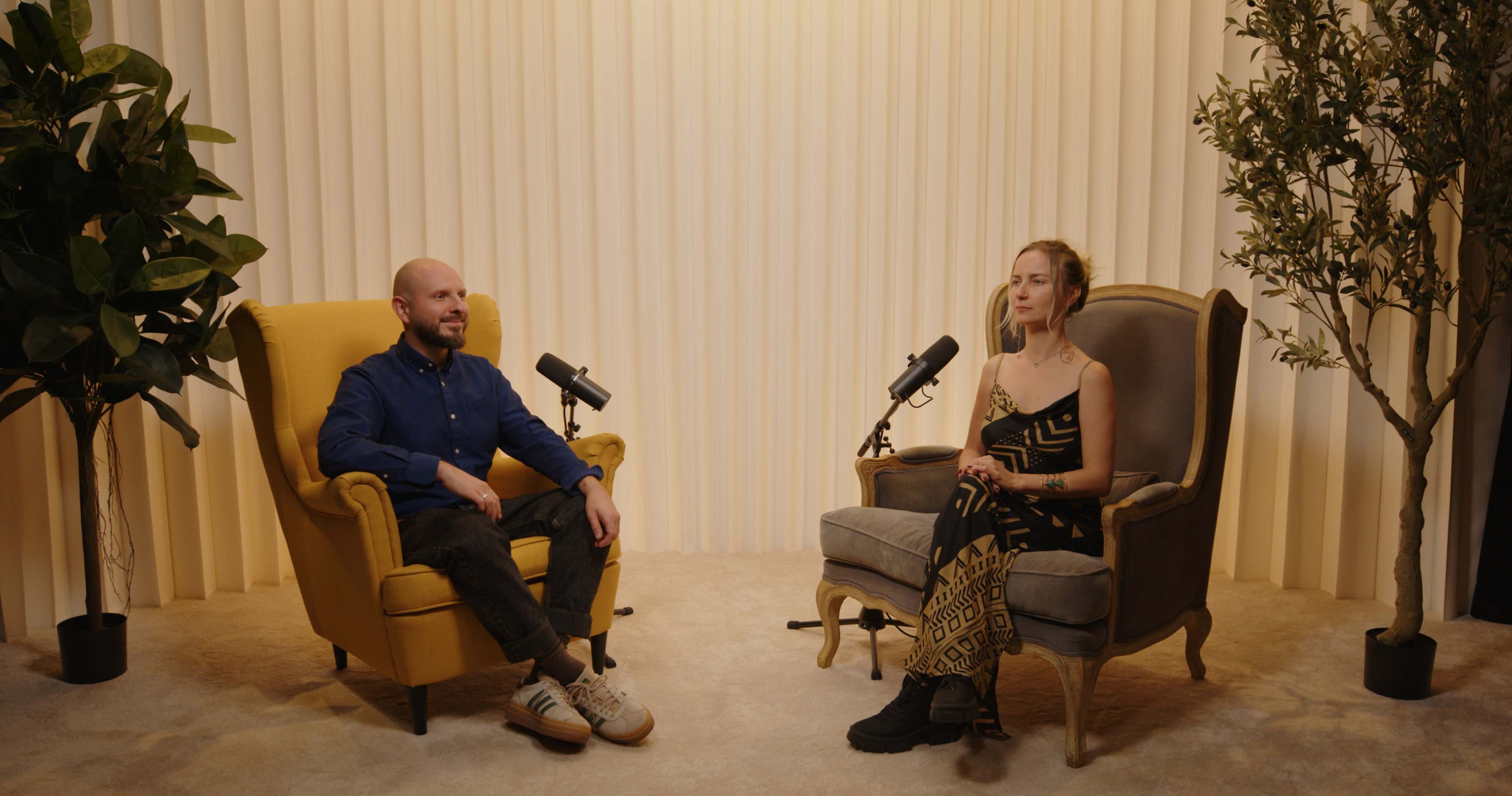 Two people are seated in armchairs facing each other, positioned under two microphones, in front of a backdrop of vertical fabric drapes.
