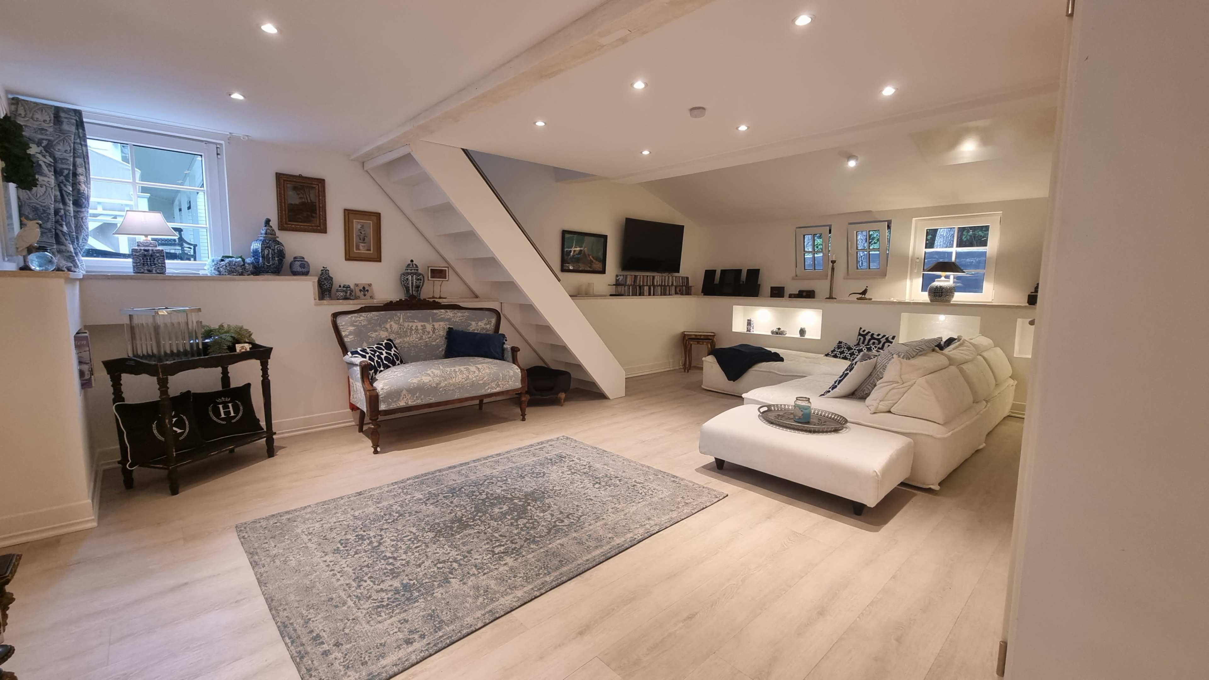 The image shows a modern, well-lit living room with a sofa, a chaise lounge, and a decorative rug on a wooden floor.