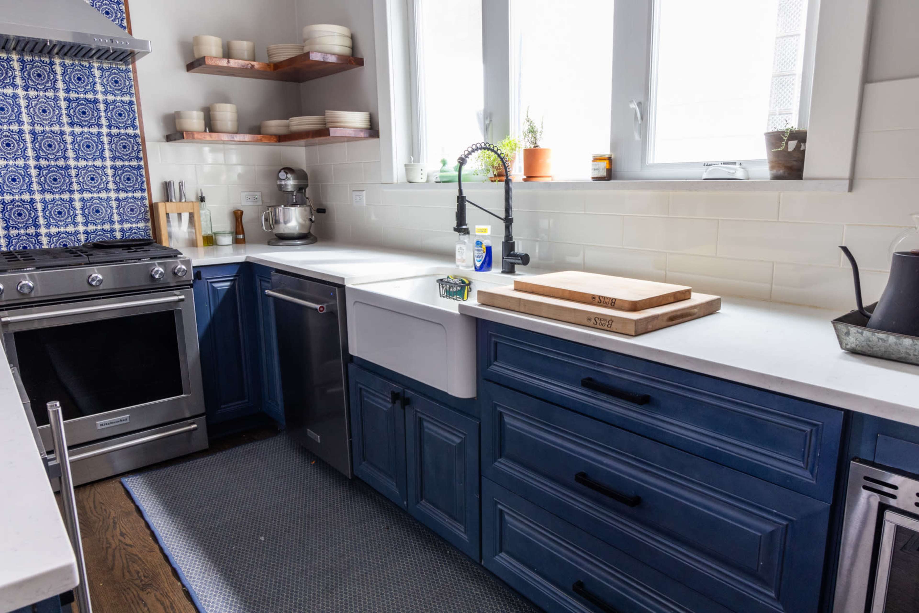 A modern kitchen with blue cabinets, a farmhouse sink, stainless steel appliances, and open shelves displaying white plates.