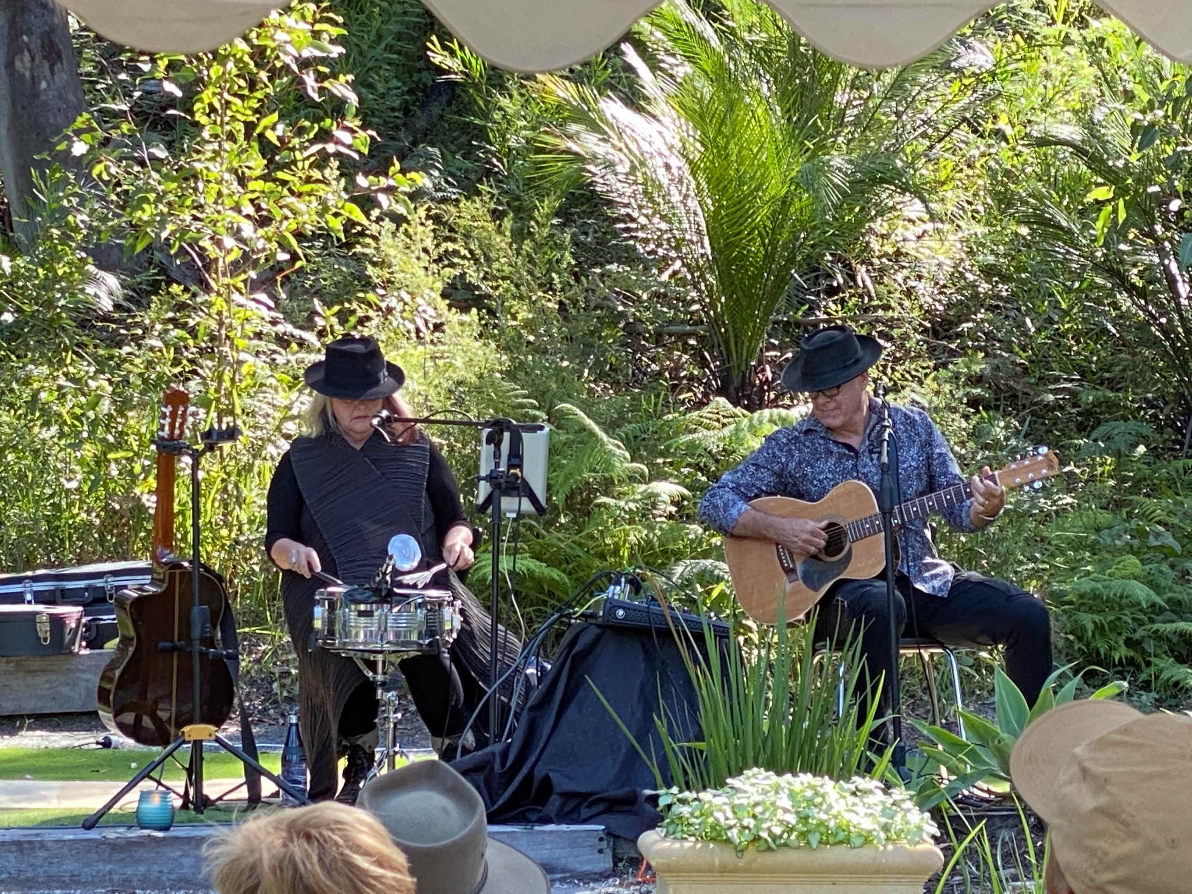 Two musicians perform outdoors with a guitar and percussion in a lush garden setting.