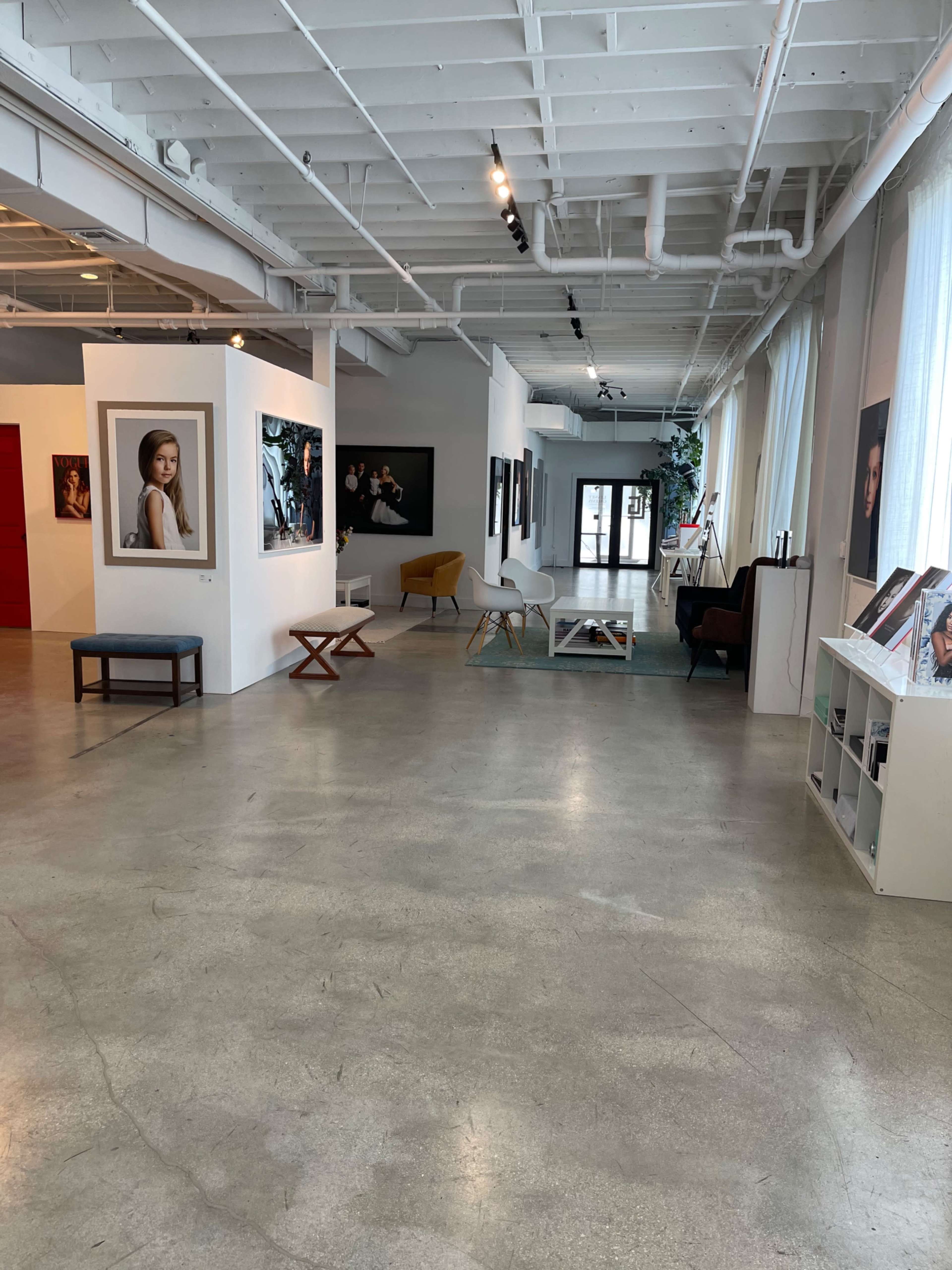 The image shows an art gallery interior with large photographs on the walls, a few seating areas, and natural light filtering in through large windows.