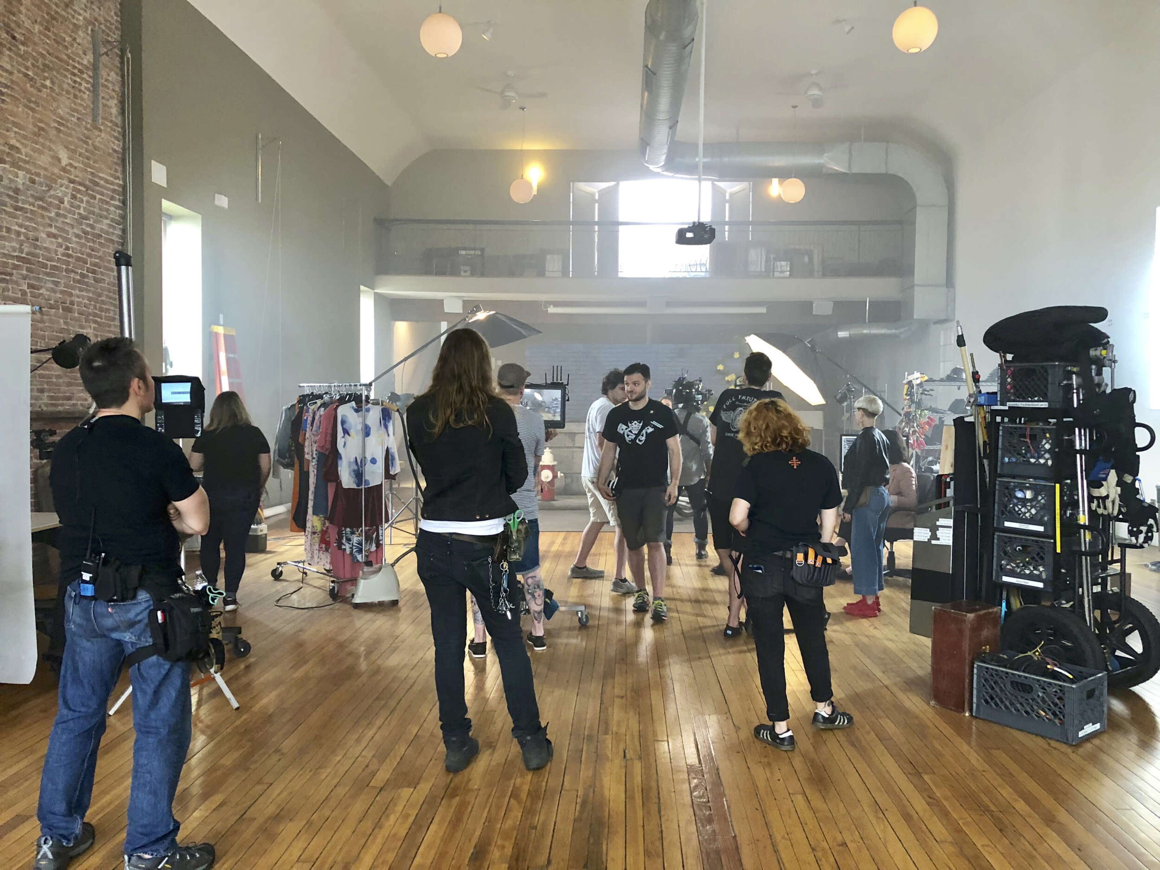 A group of people are preparing for a photoshoot in a spacious, well-lit studio filled with clothing racks and various equipment.