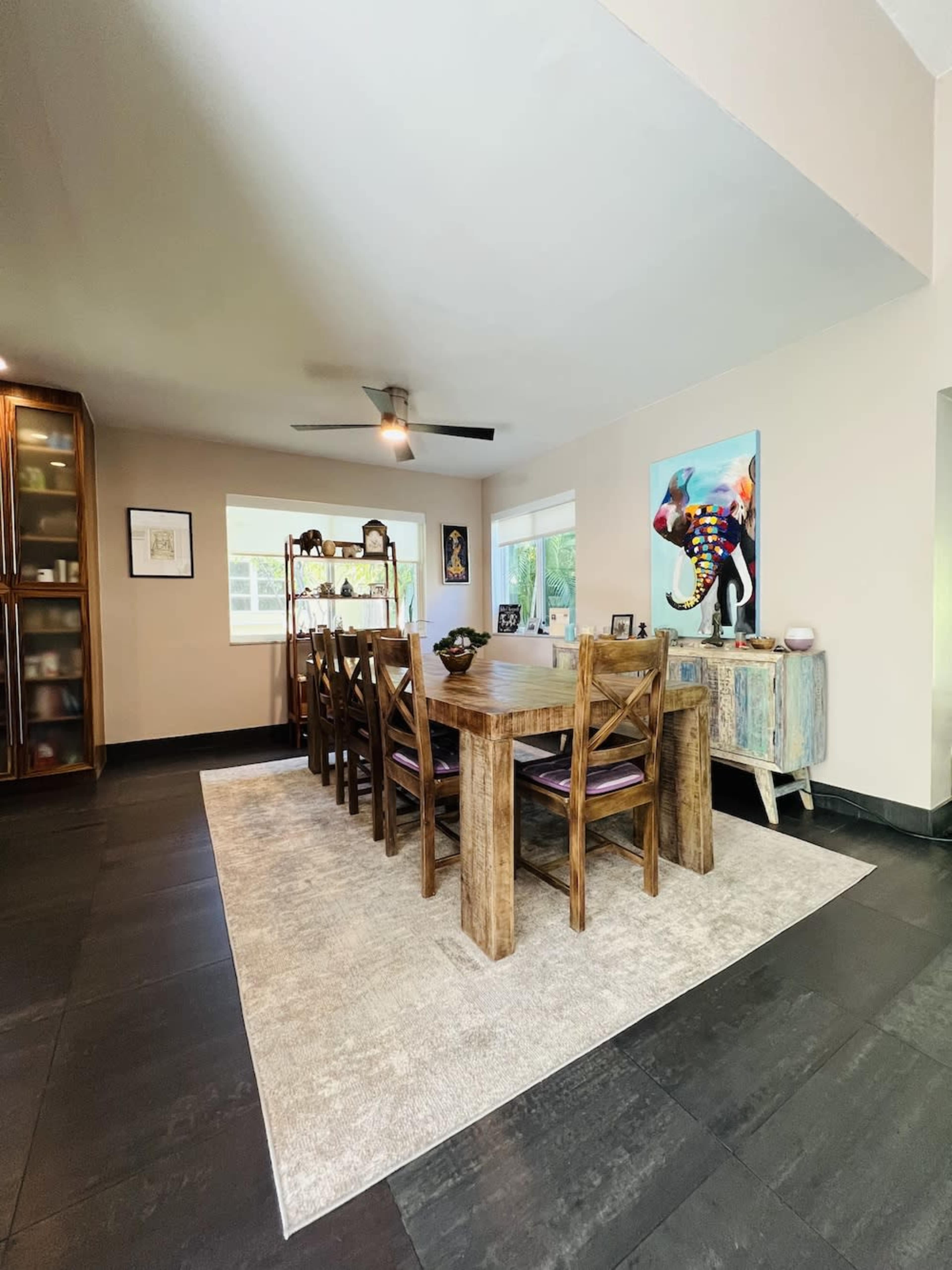 A rectangular wooden dining table is surrounded by eight chairs, set on a light gray rug in a well-lit room featuring a sideboard and artwork on the walls.