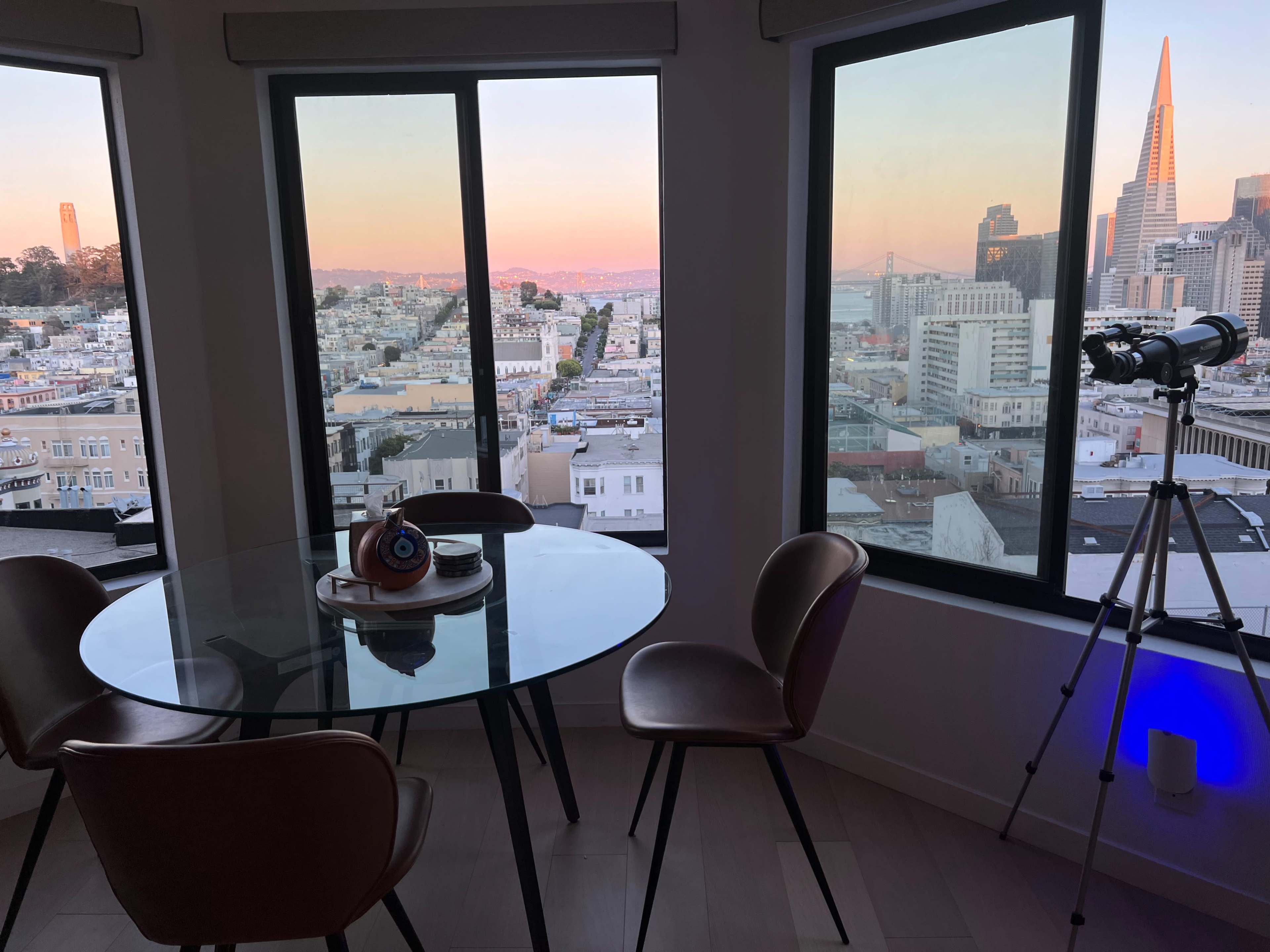 A glass dining table with three chairs is positioned near large windows, showing a view of San Francisco's skyline at sunset, with a telescope set up next to the window.