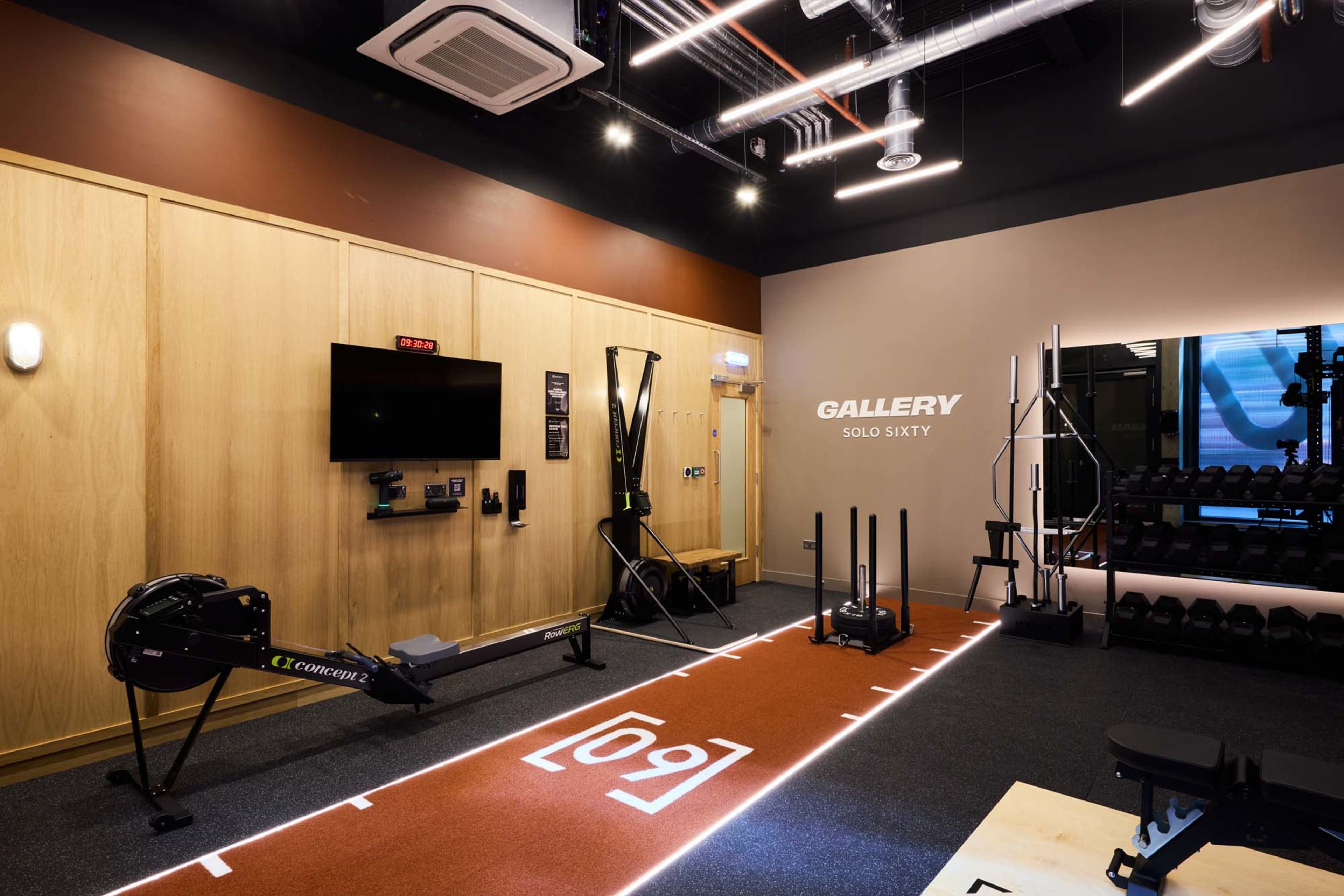 The image shows a modern fitness studio featuring exercise equipment such as rowing machines, dumbbells, and a sled on a carpeted area marked with a lane design.