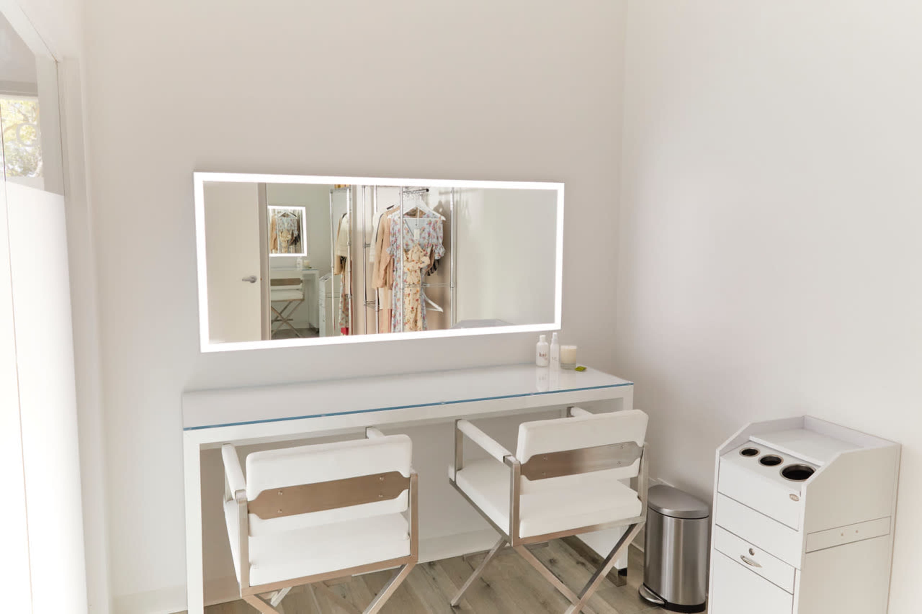 A clean, well-lit dressing area with two white chairs in front of a mirror and a clothing rack in the background.