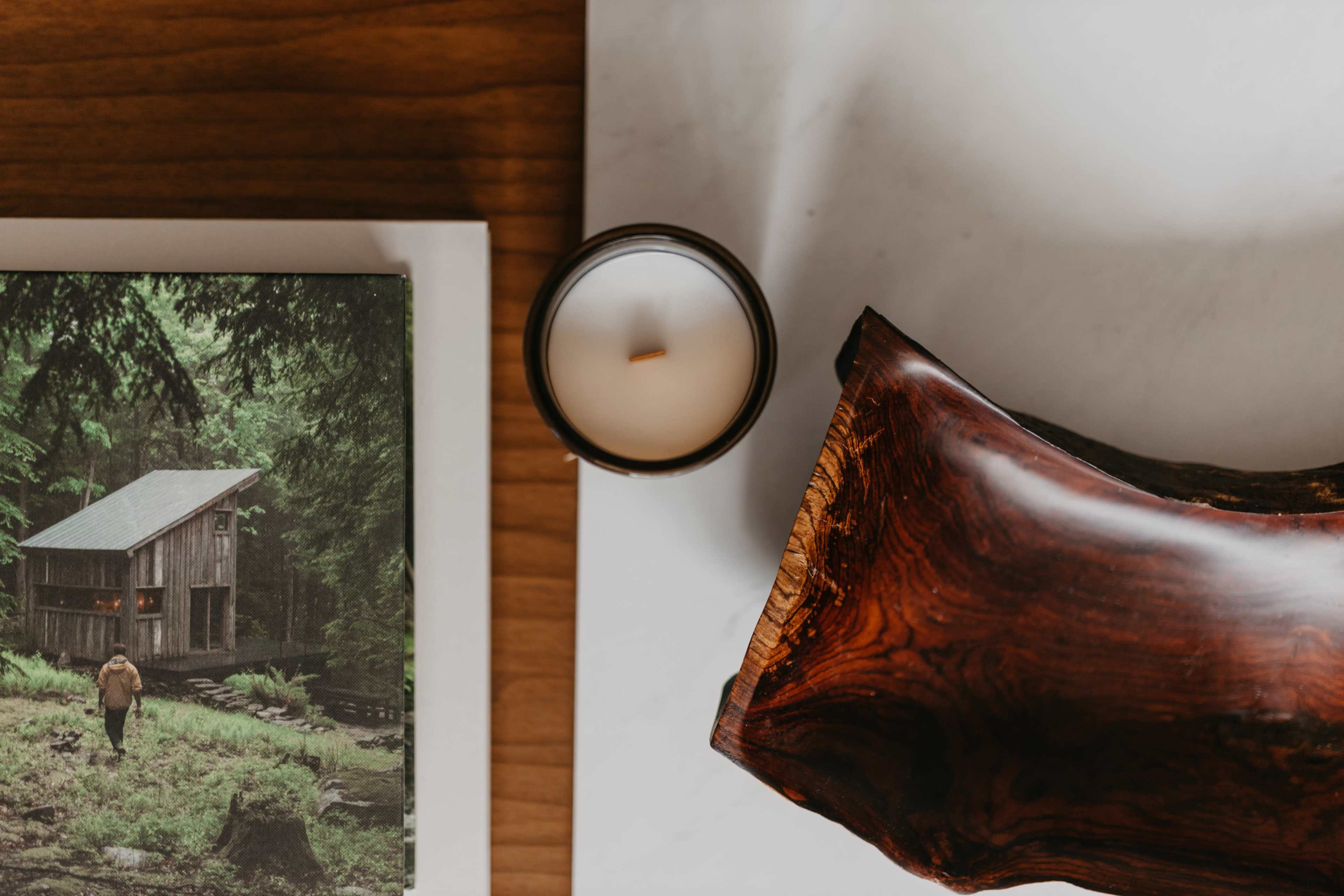 A wooden surface holds a candle, a piece of polished wood, and a book with a scenic image of a cabin in a forest.