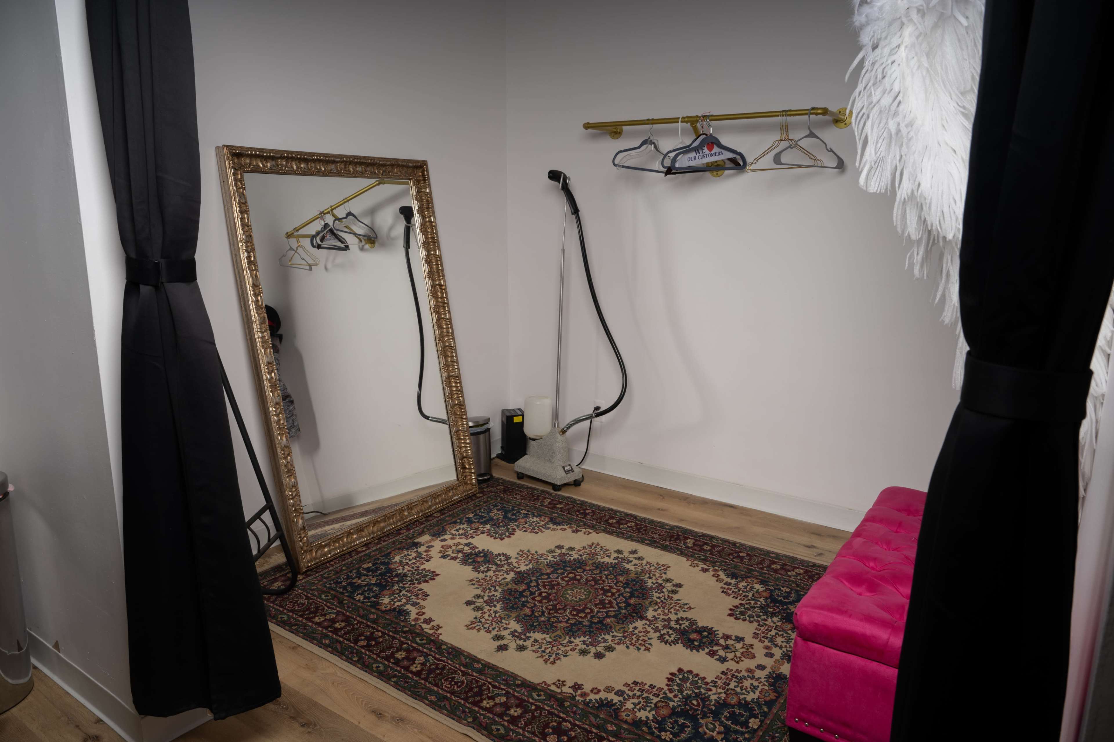 A fitting room with a large mirror, a pink bench, clothing hangers on a wall, and a garment steamer on the floor.