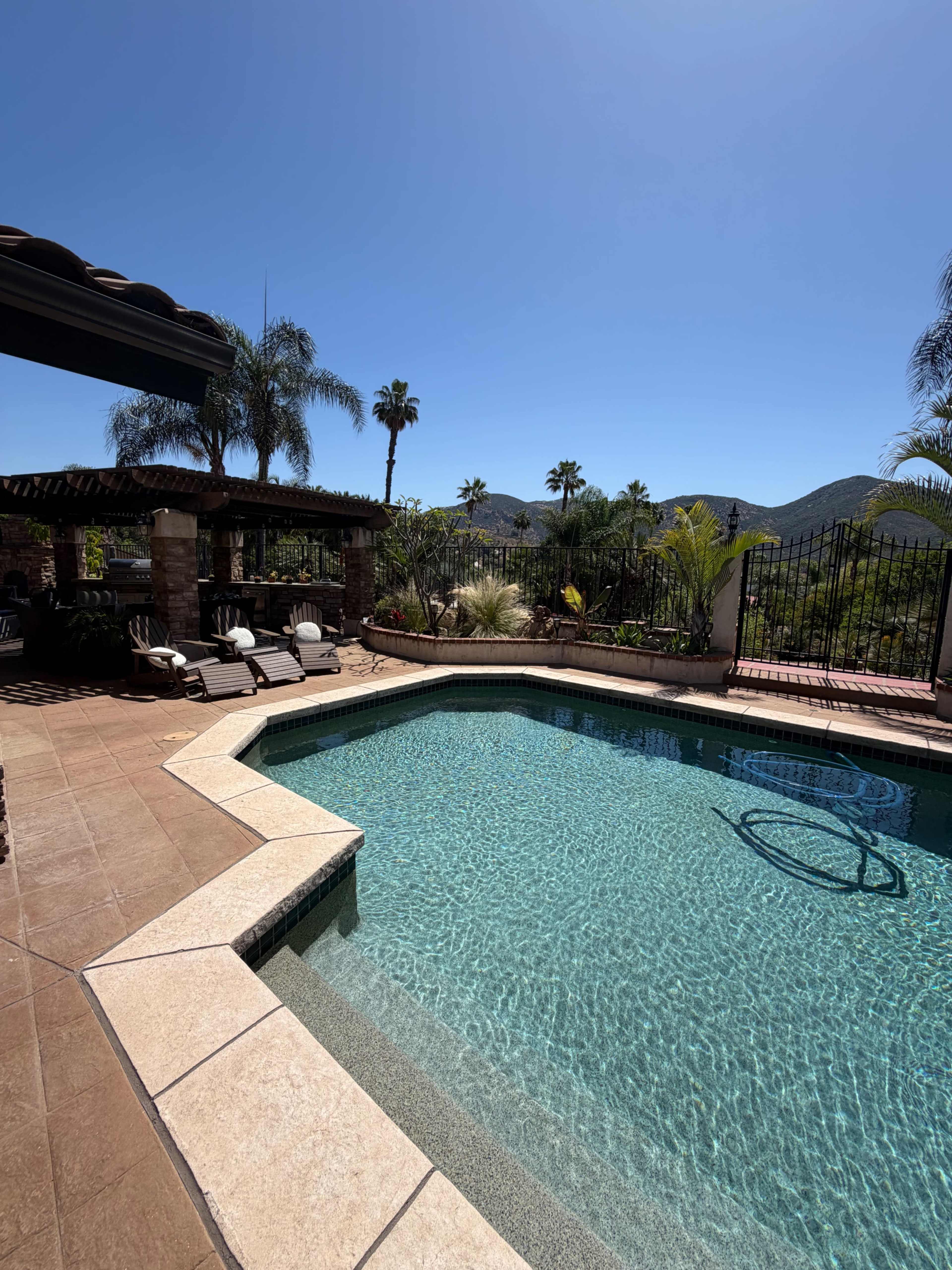 A clear swimming pool is surrounded by a stone deck and palm trees, with mountains visible in the background under a blue sky.