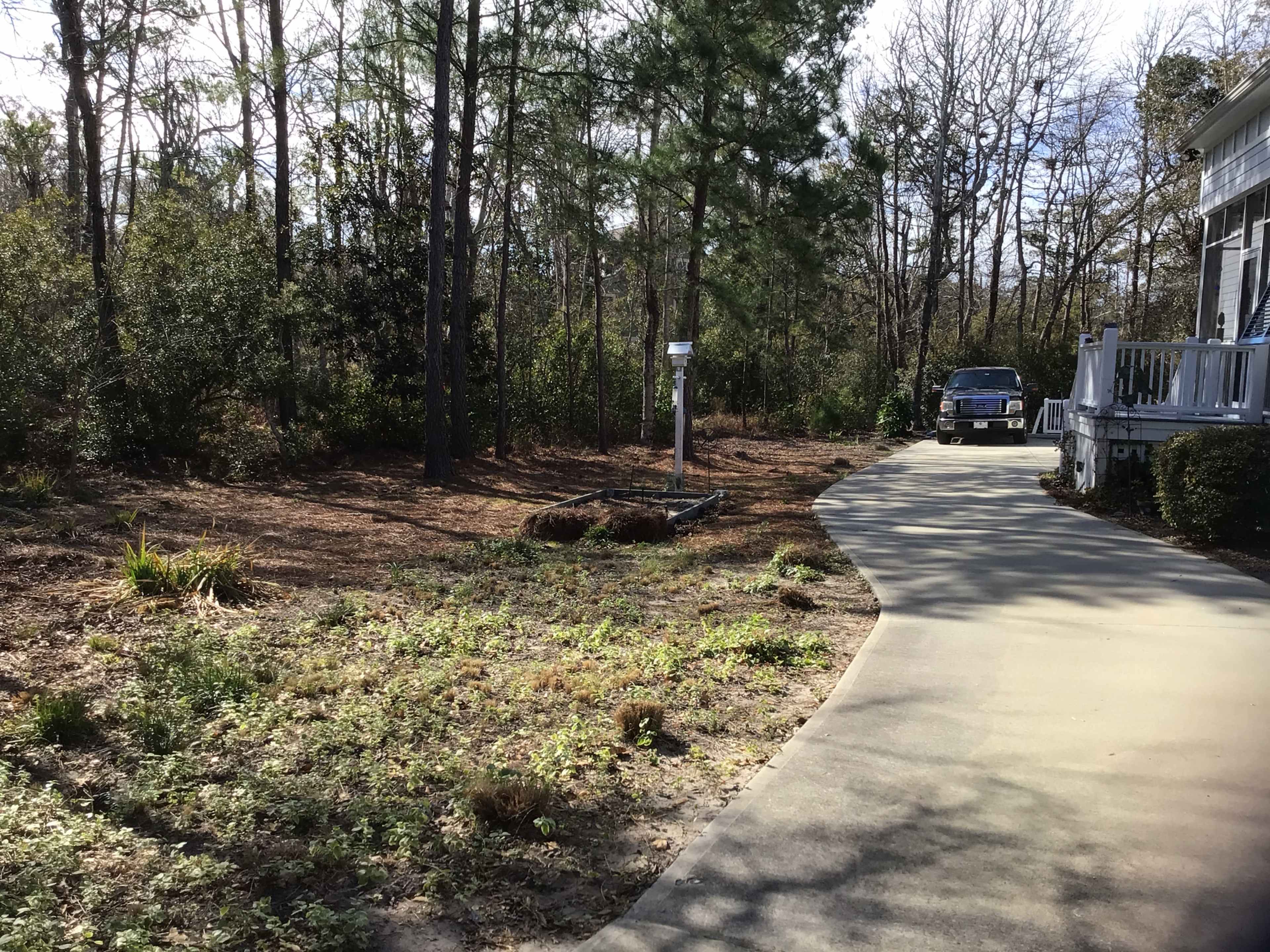 A paved driveway leads past a sparse garden area to a house surrounded by trees.