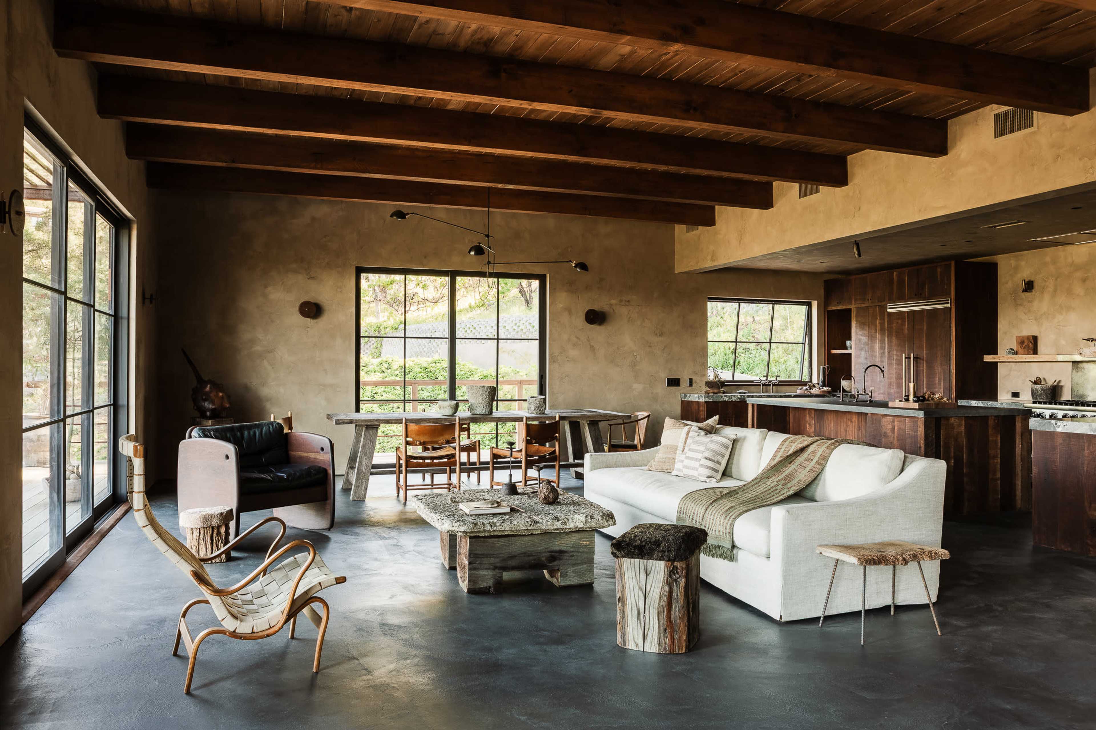 The image shows a modern living area featuring a white sofa, a wooden coffee table, and large windows that overlook a green outdoor space.