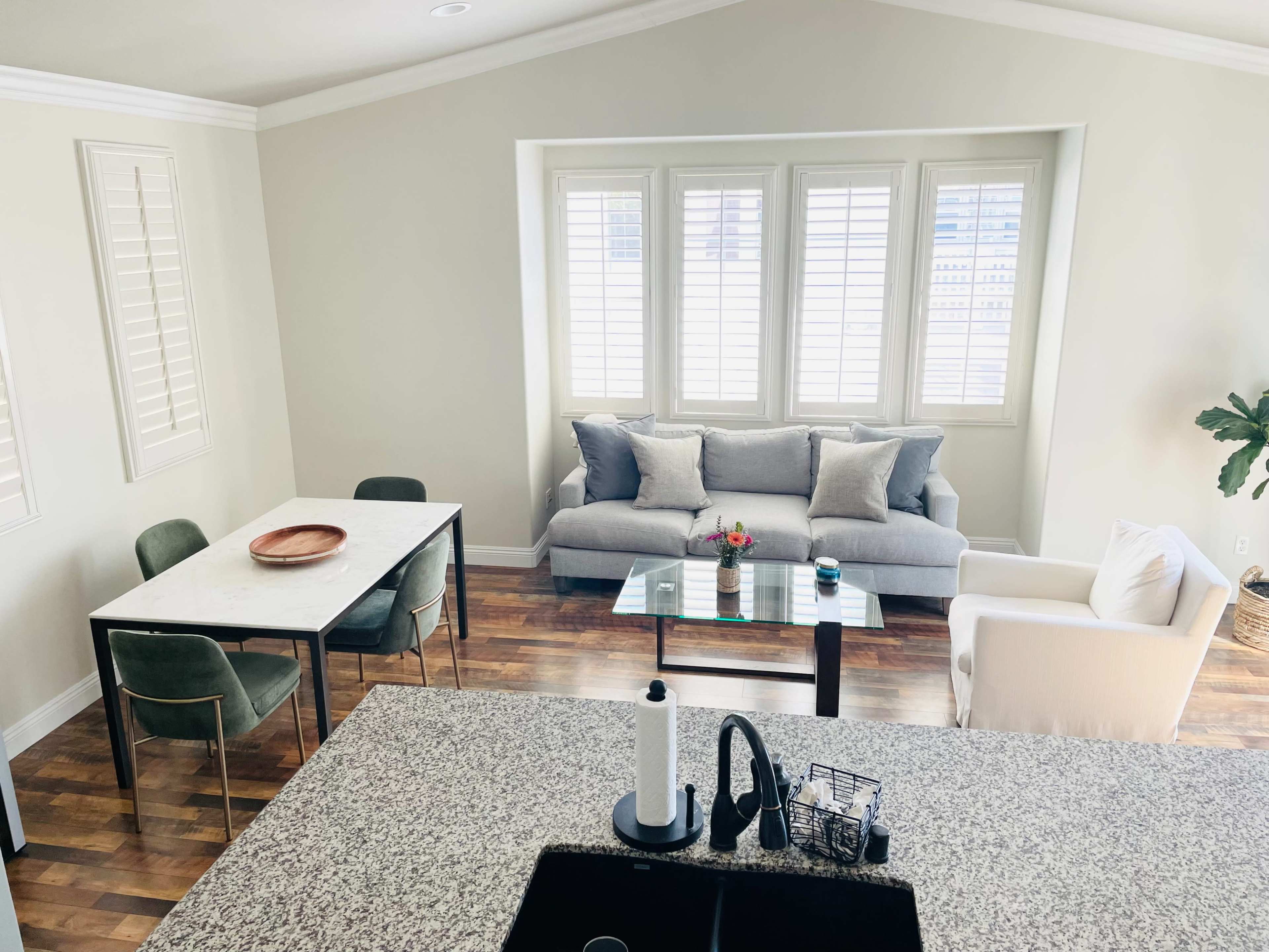 A modern living space features a dining table, a sofa with decorative cushions, and large windows with shutters, complemented by wooden flooring.