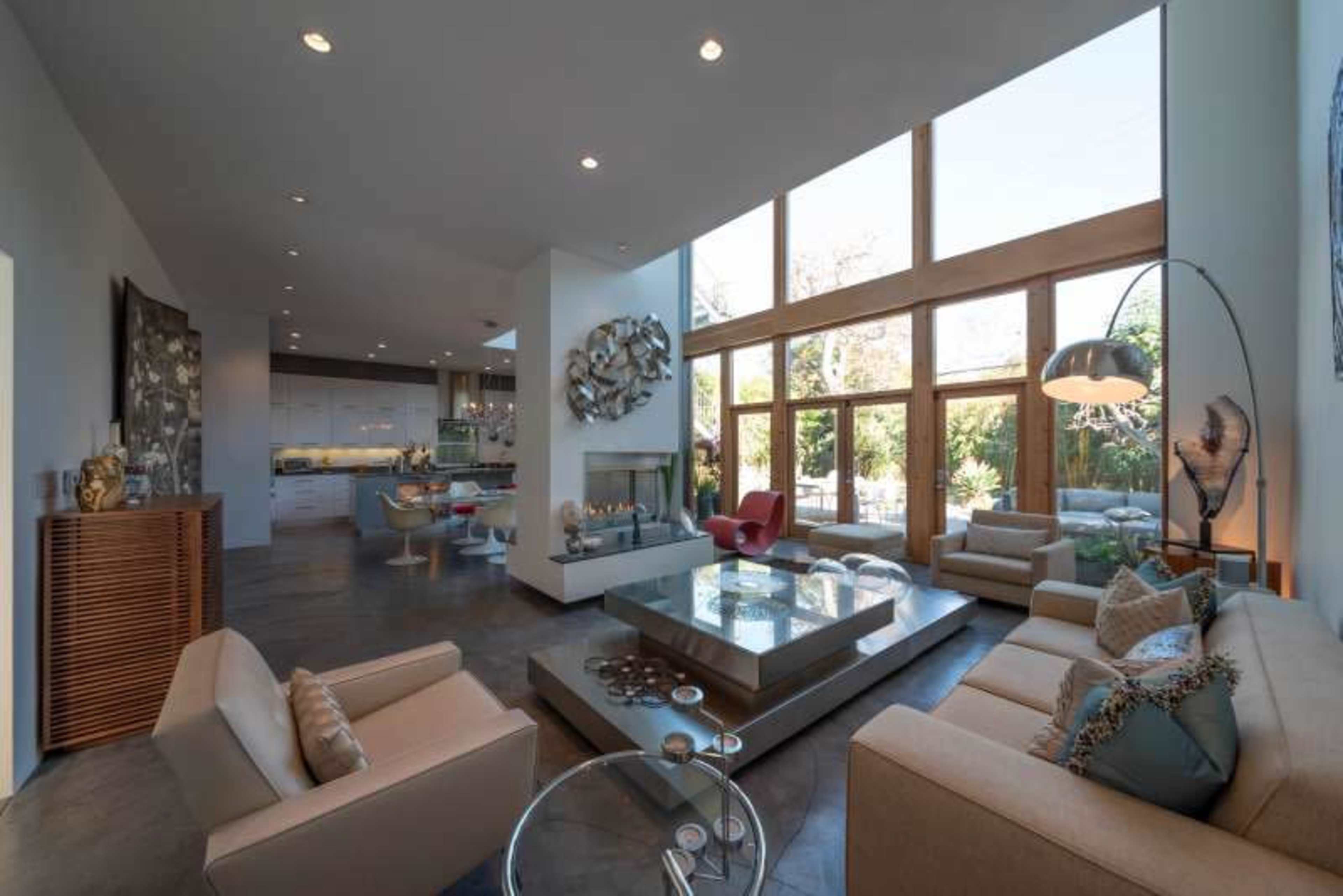 A modern living room with large windows, a central coffee table, and contemporary furniture that faces a fireplace and an open kitchen area.