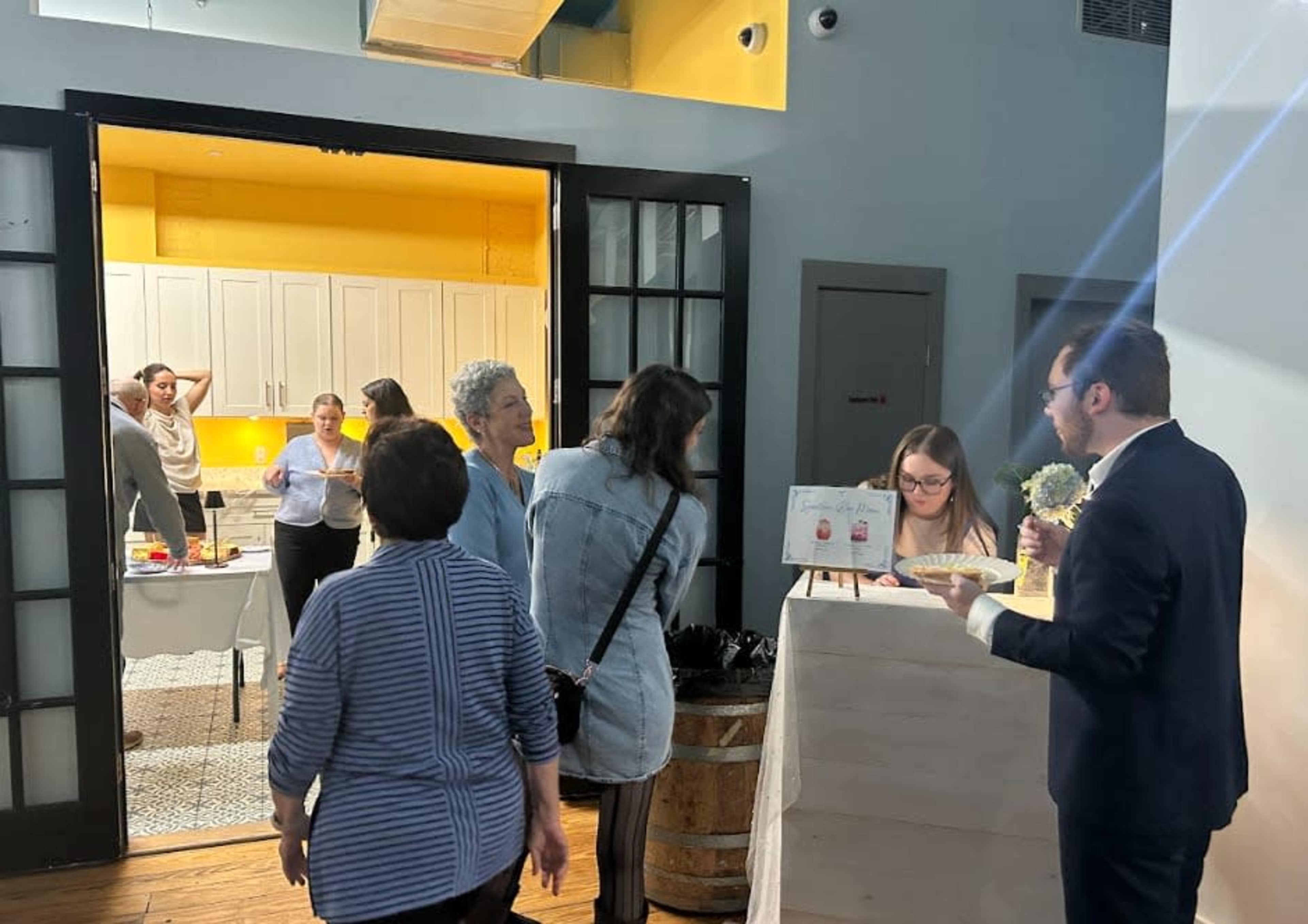 A group of people engages in conversation while a woman assists a customer at a counter in a brightly lit room.