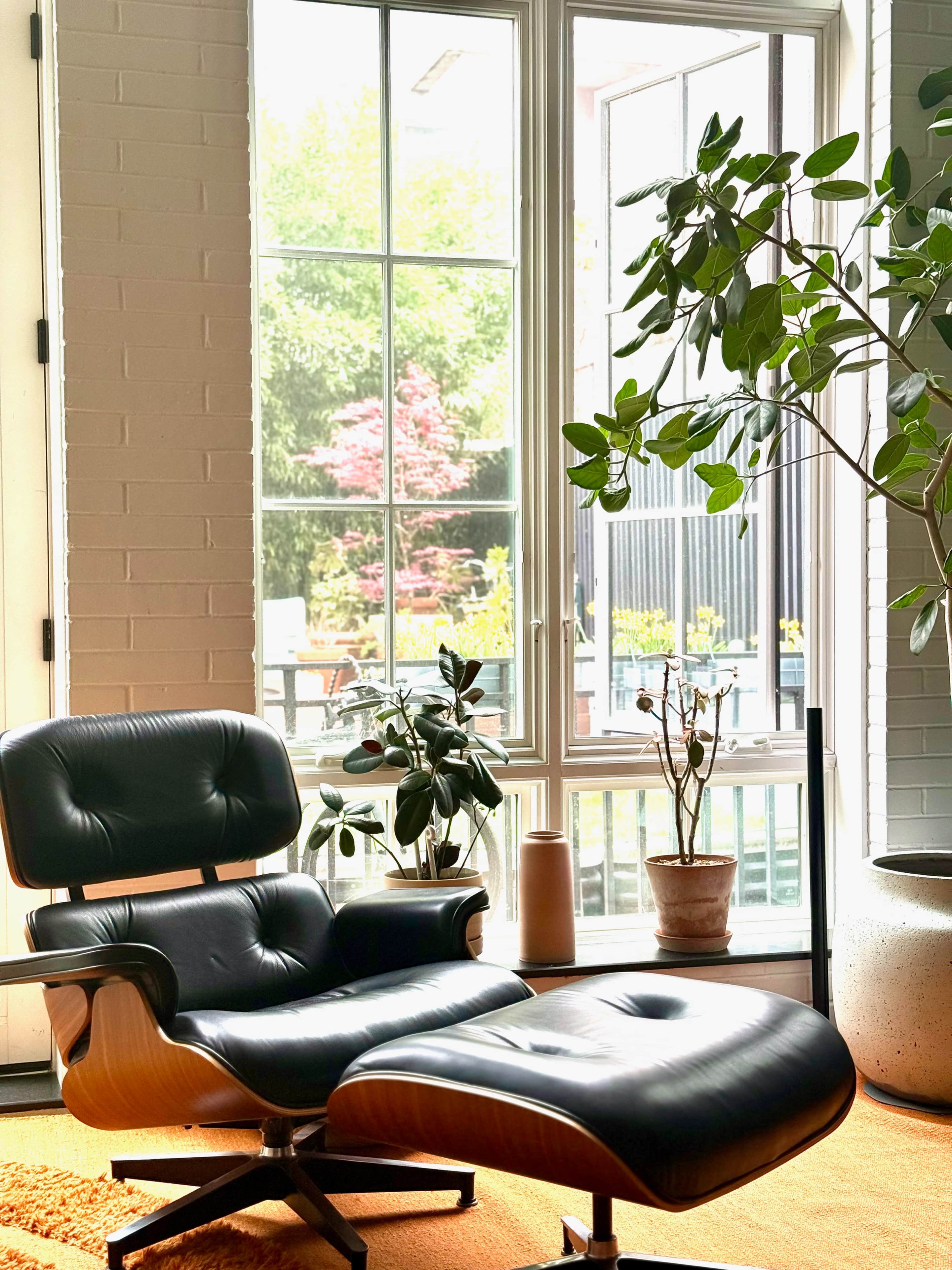 A black leather chair with an ottoman is positioned in front of large windows that overlook a garden filled with plants and trees.