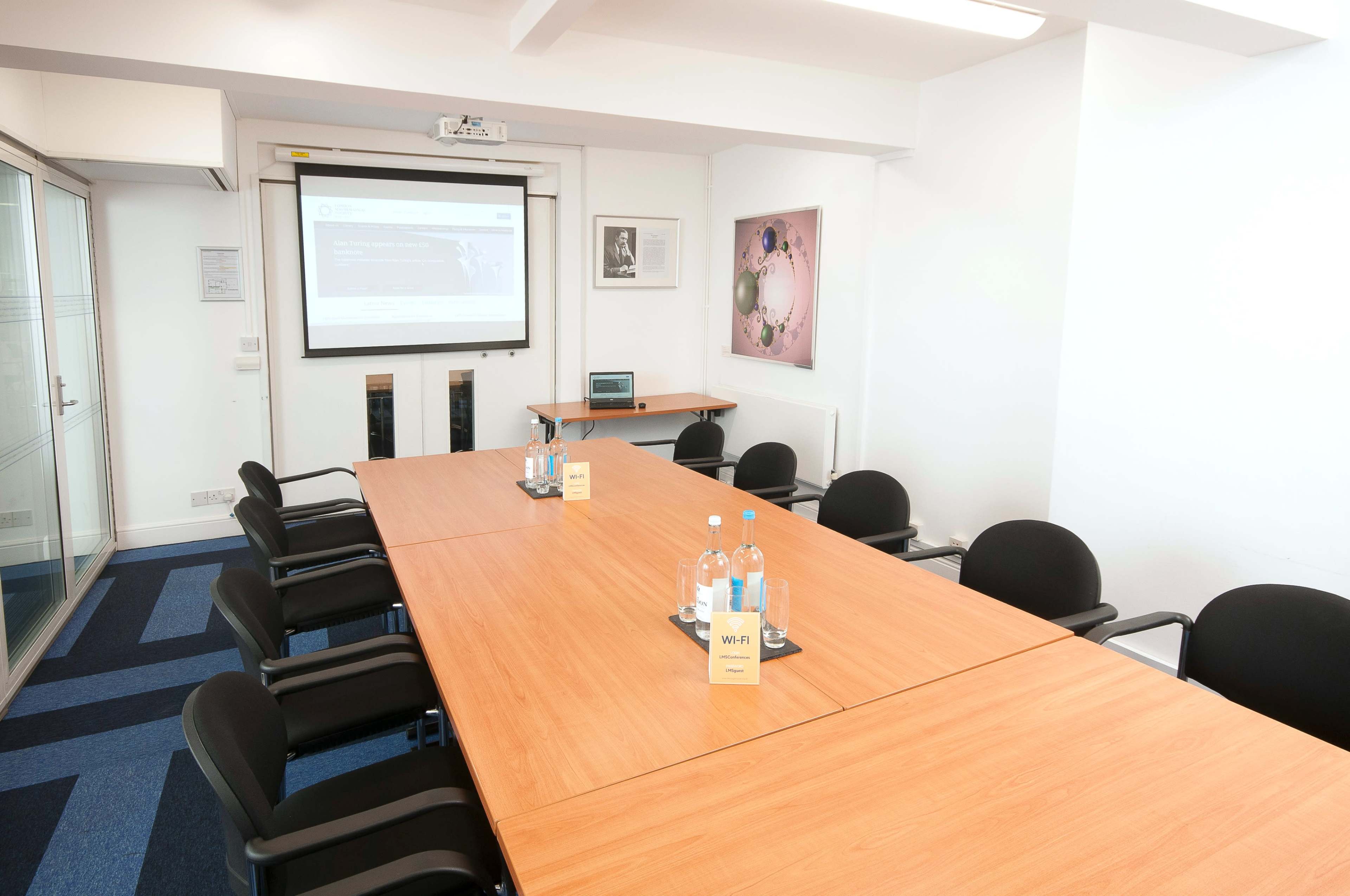 A conference room features a long wooden table surrounded by black chairs, a projector screen mounted on the wall, and bottles of water on the table.