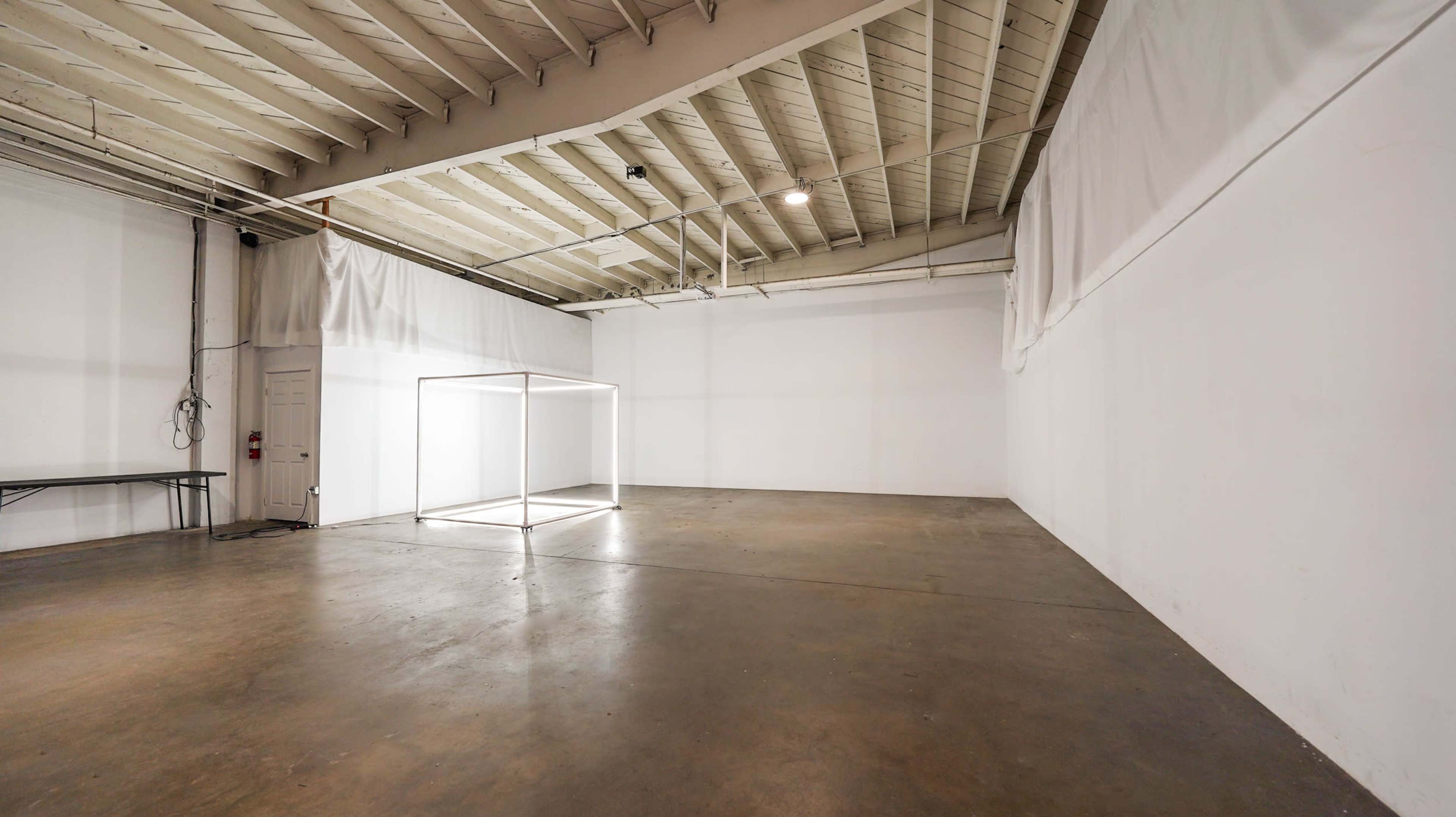 The image shows a spacious, empty room with white walls, a concrete floor, and a lit square structure in the center.