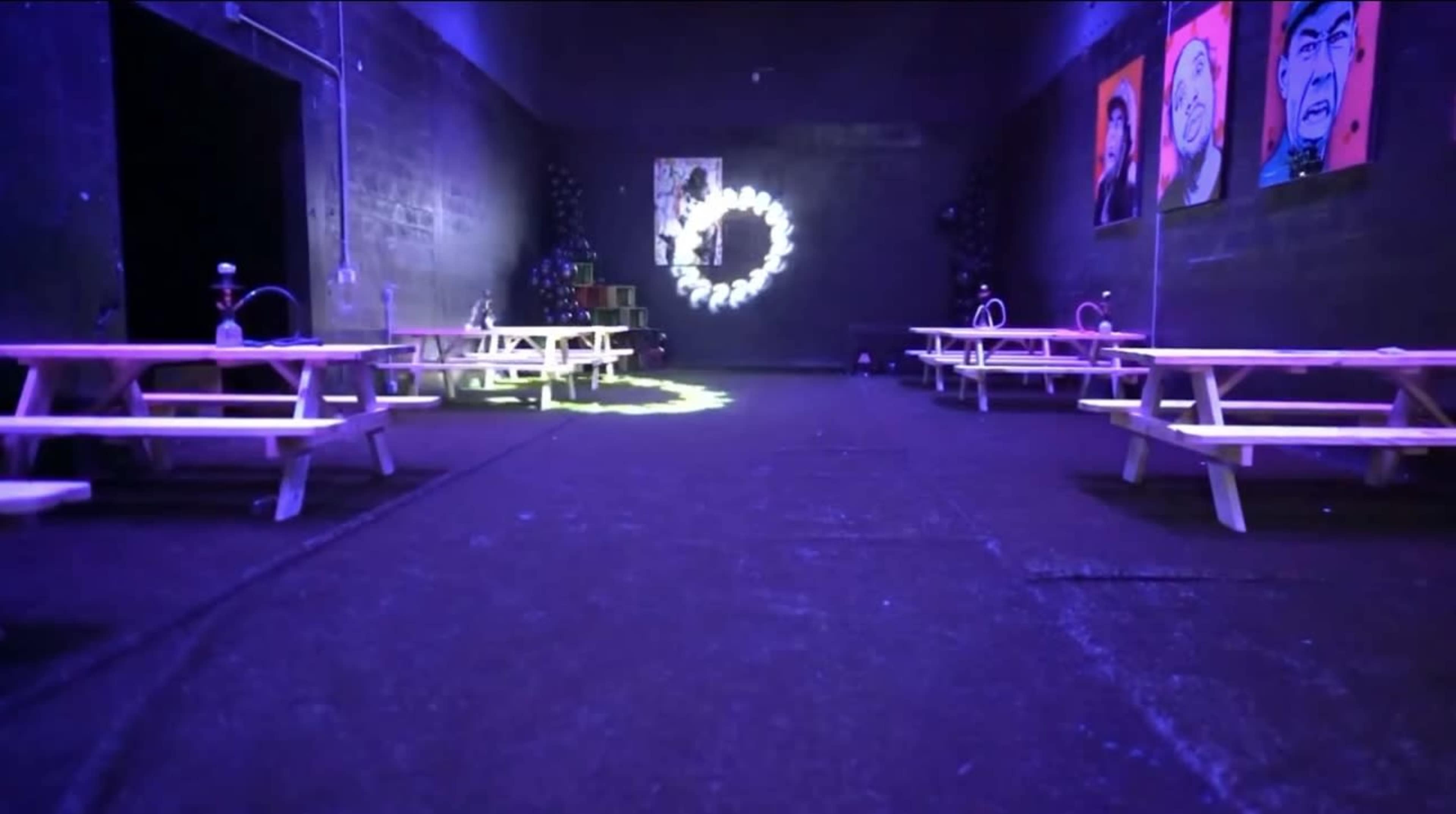 The image shows a dimly lit indoor space with wooden picnic tables arranged along the sides and colorful artwork on the walls.