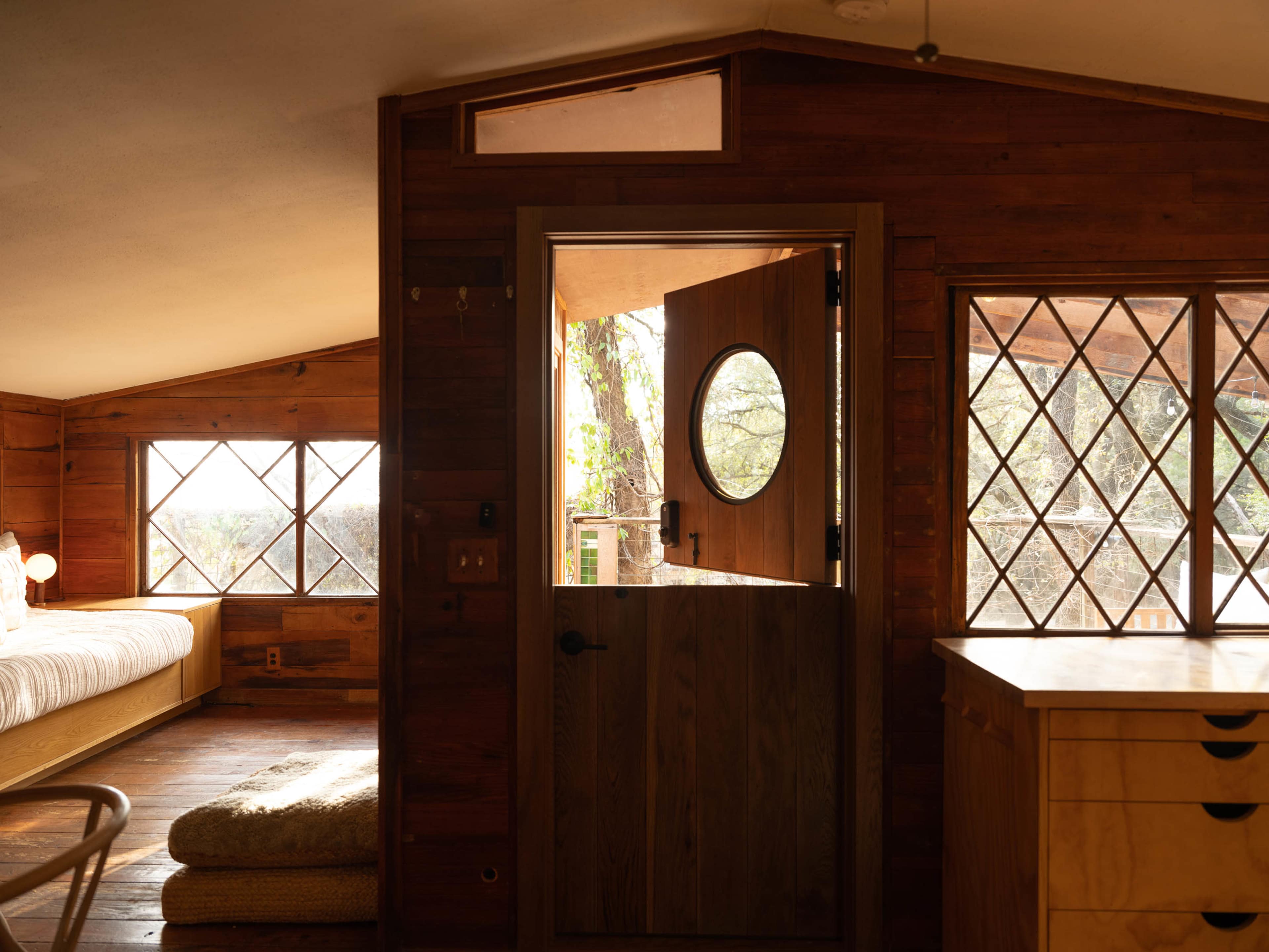 A cozy wooden interior with a circular windowed door leading to an outdoor area, framed by diamond-patterned windows.