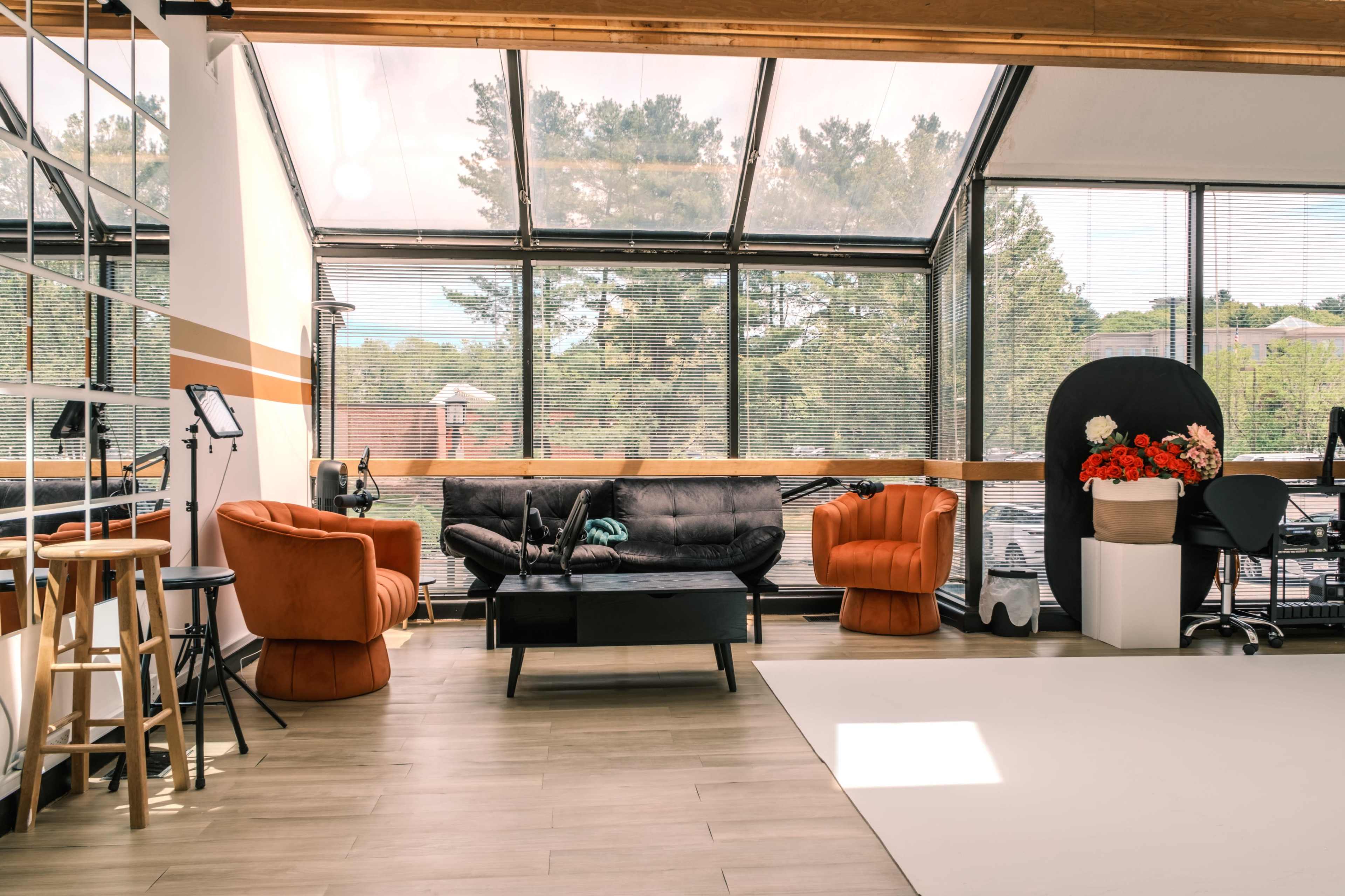 The image shows a modern office space with large windows, featuring two distinct seating areas, a black couch, a coffee table, and a backdrop for photography.