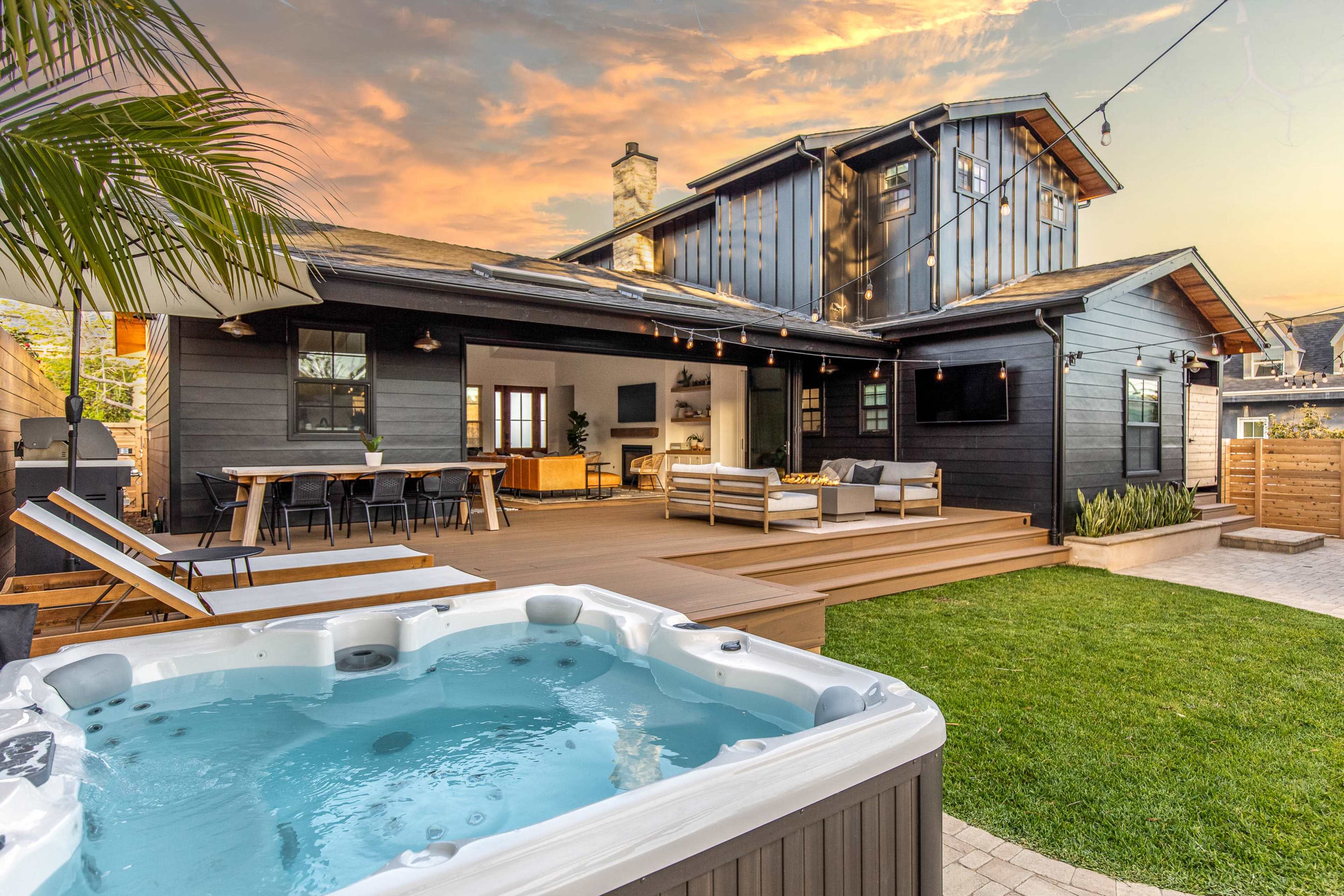 A modern backyard features a hot tub in the foreground, a spacious deck with outdoor furniture, and a black and wood-finished house under a colorful sunset.