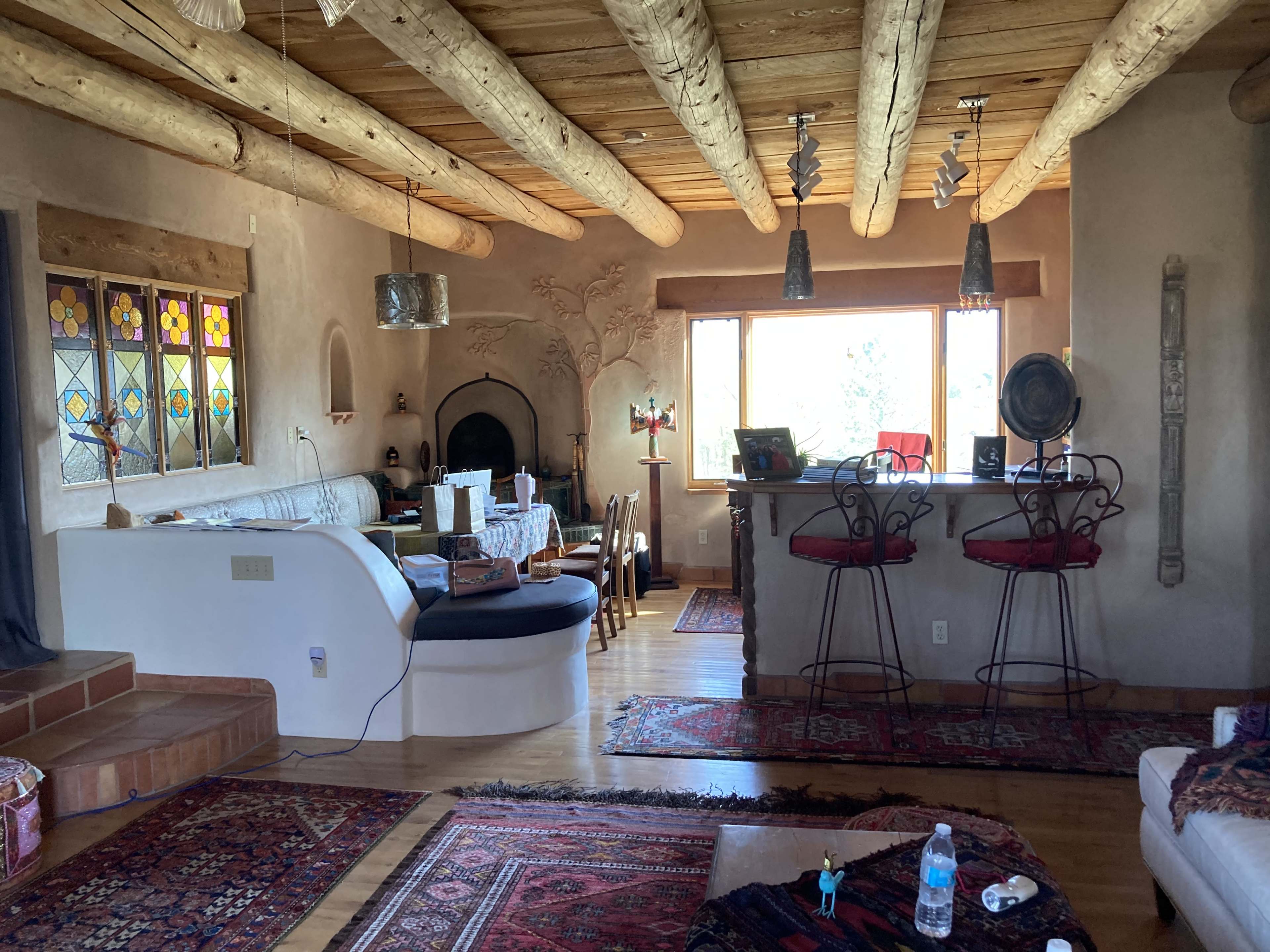A rustic living room features wooden beams, stained glass windows, and a dining area with a table, surrounded by decorative rugs and a bar with tall stools.