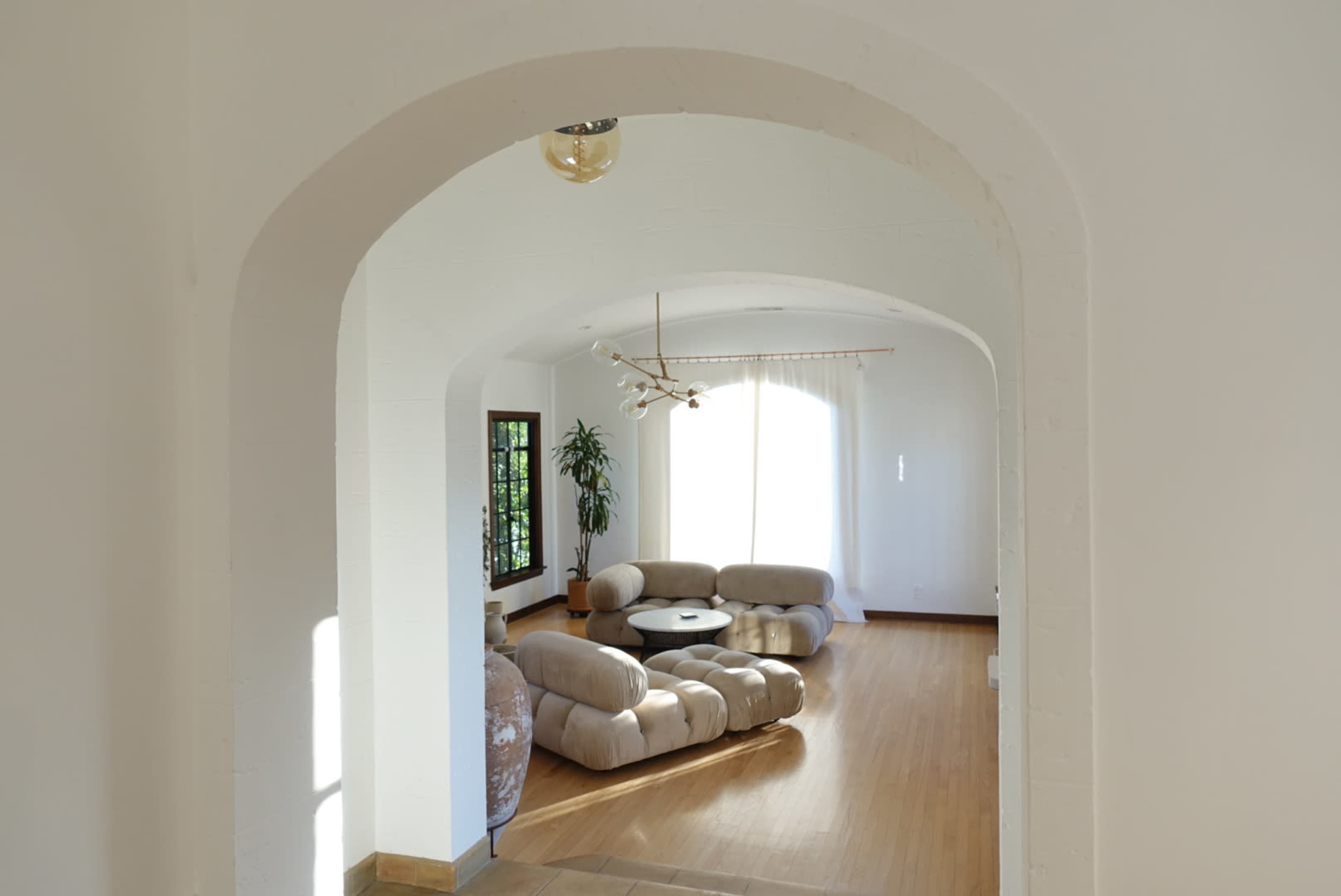A spacious living room features a curved, beige sectional sofa, a round black coffee table, and large windows with natural light.