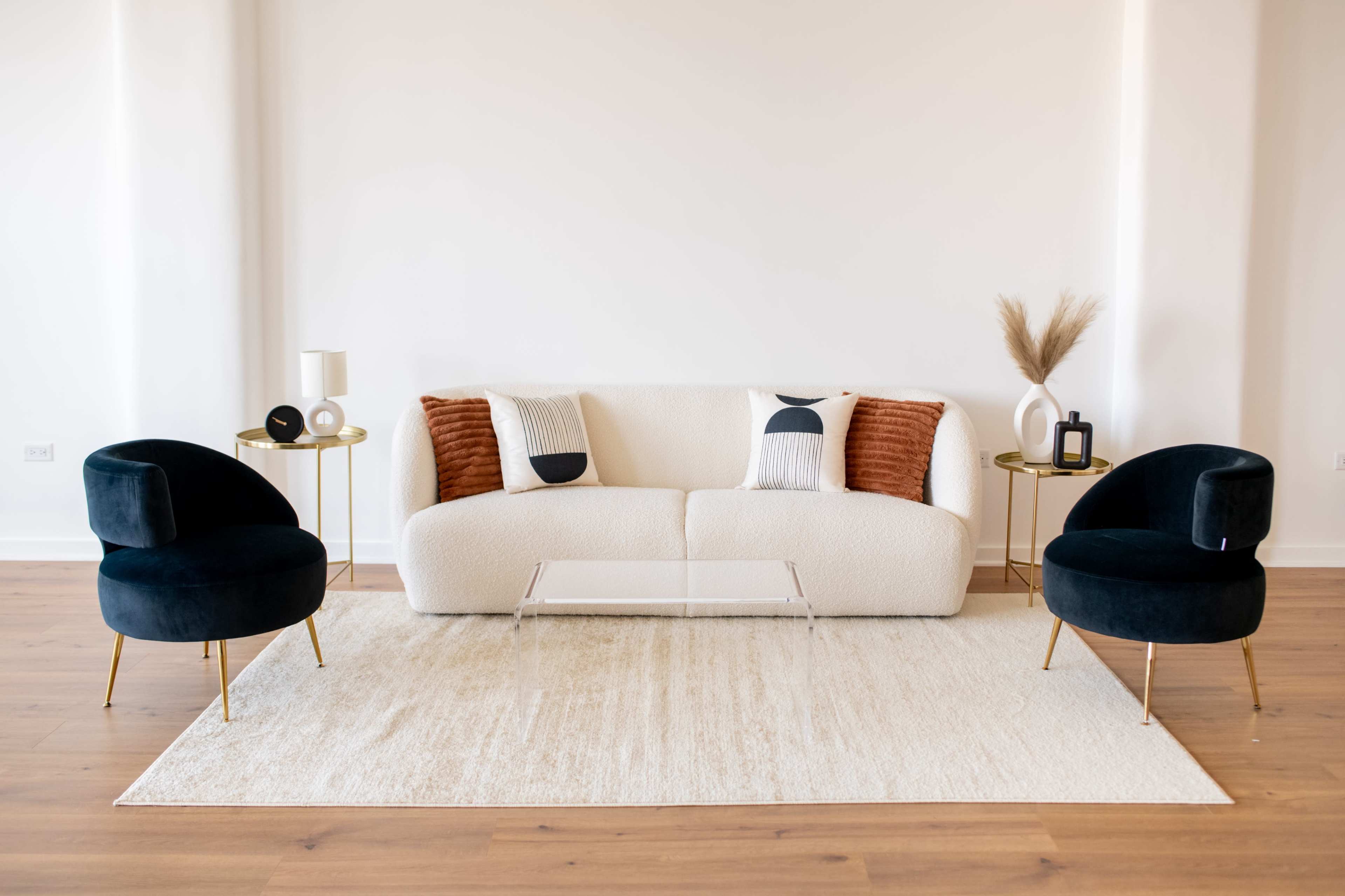 A modern living room featuring a white sofa with orange and black accents, two dark chairs, a glass coffee table, and decorative elements on side tables.