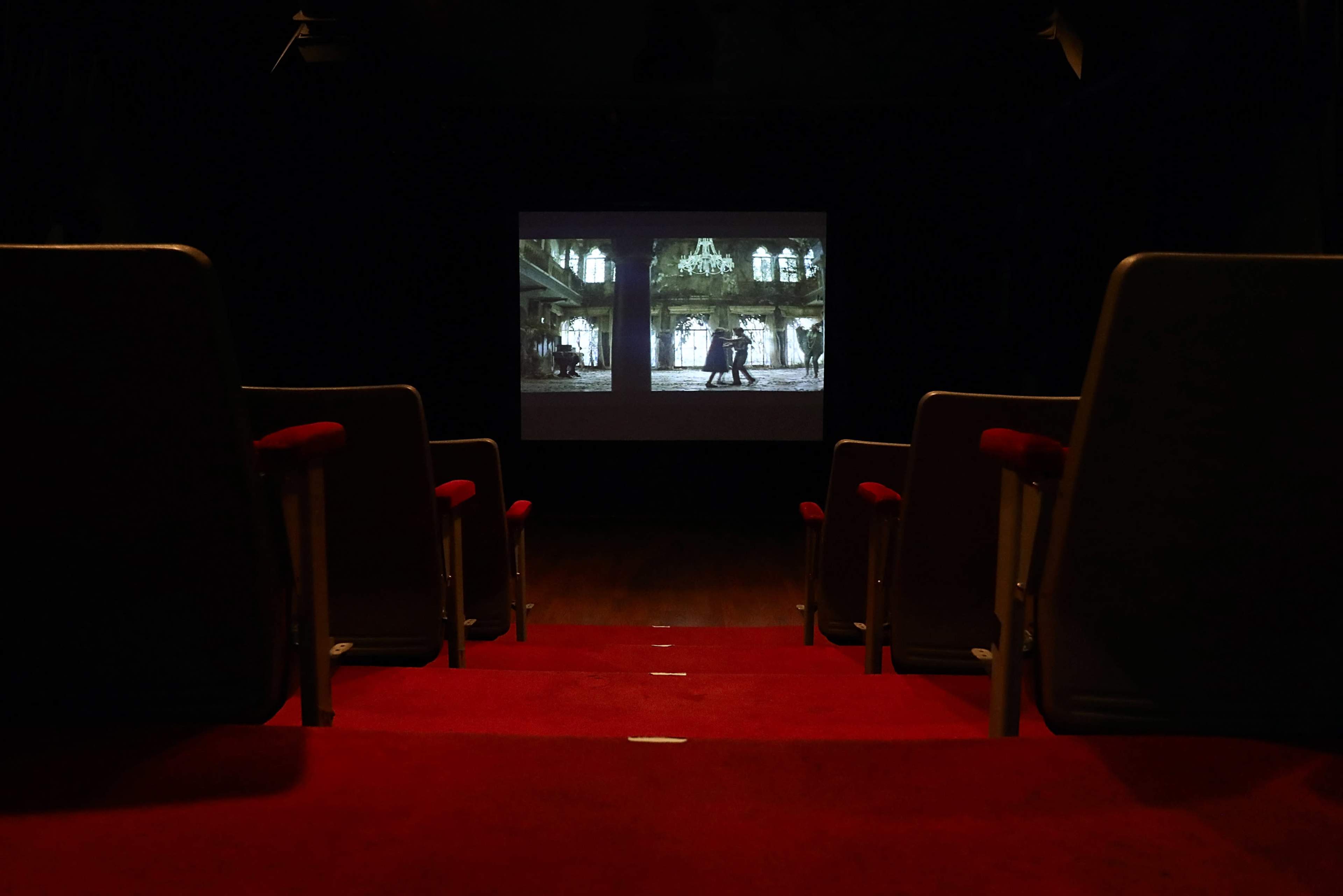 The image shows a row of empty red theater seats facing a large projection screen displaying a film scene.