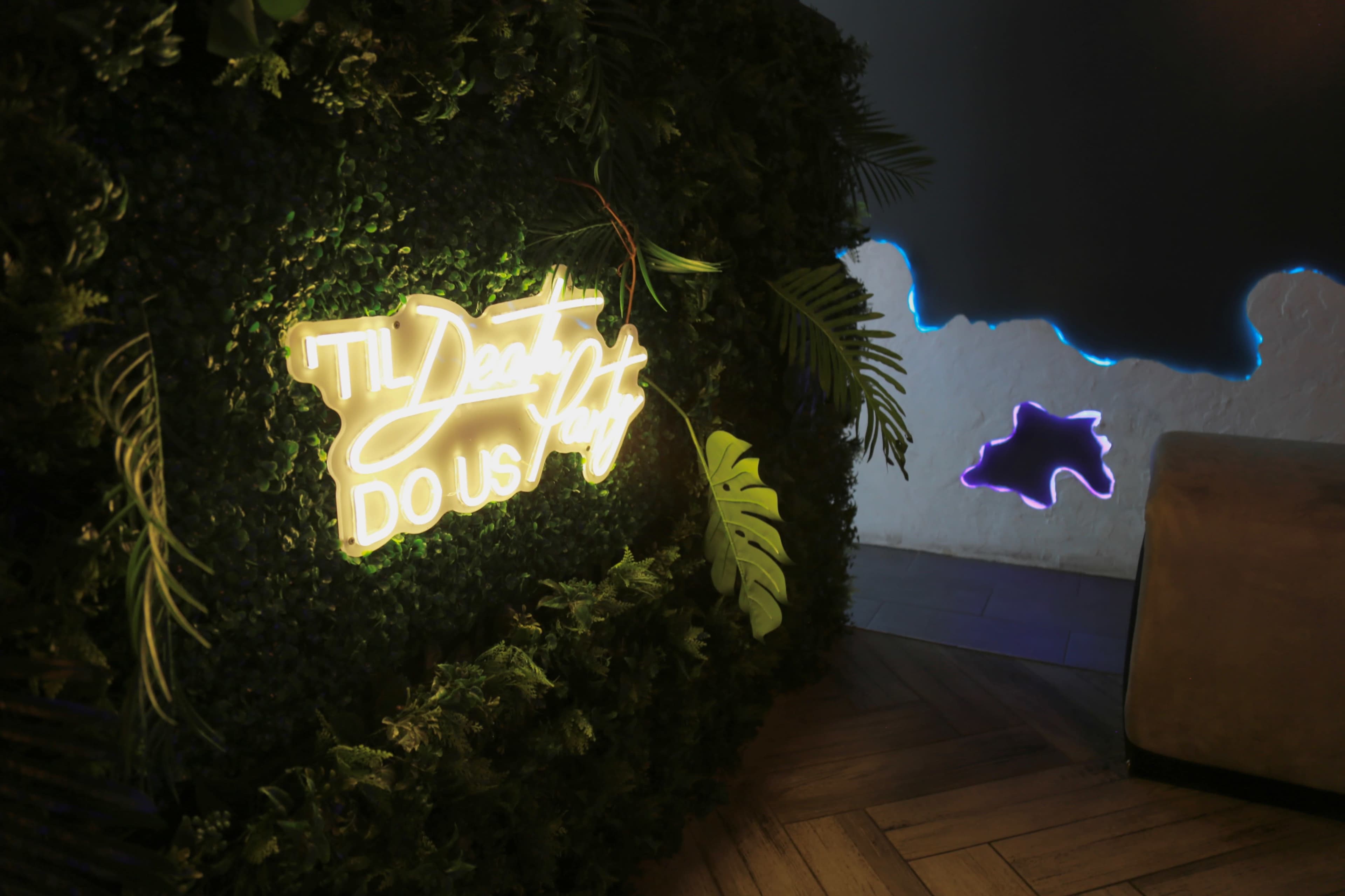 A neon sign reading "I'll Dead, Do Us Part" is surrounded by greenery on a wall in a dimly lit space.